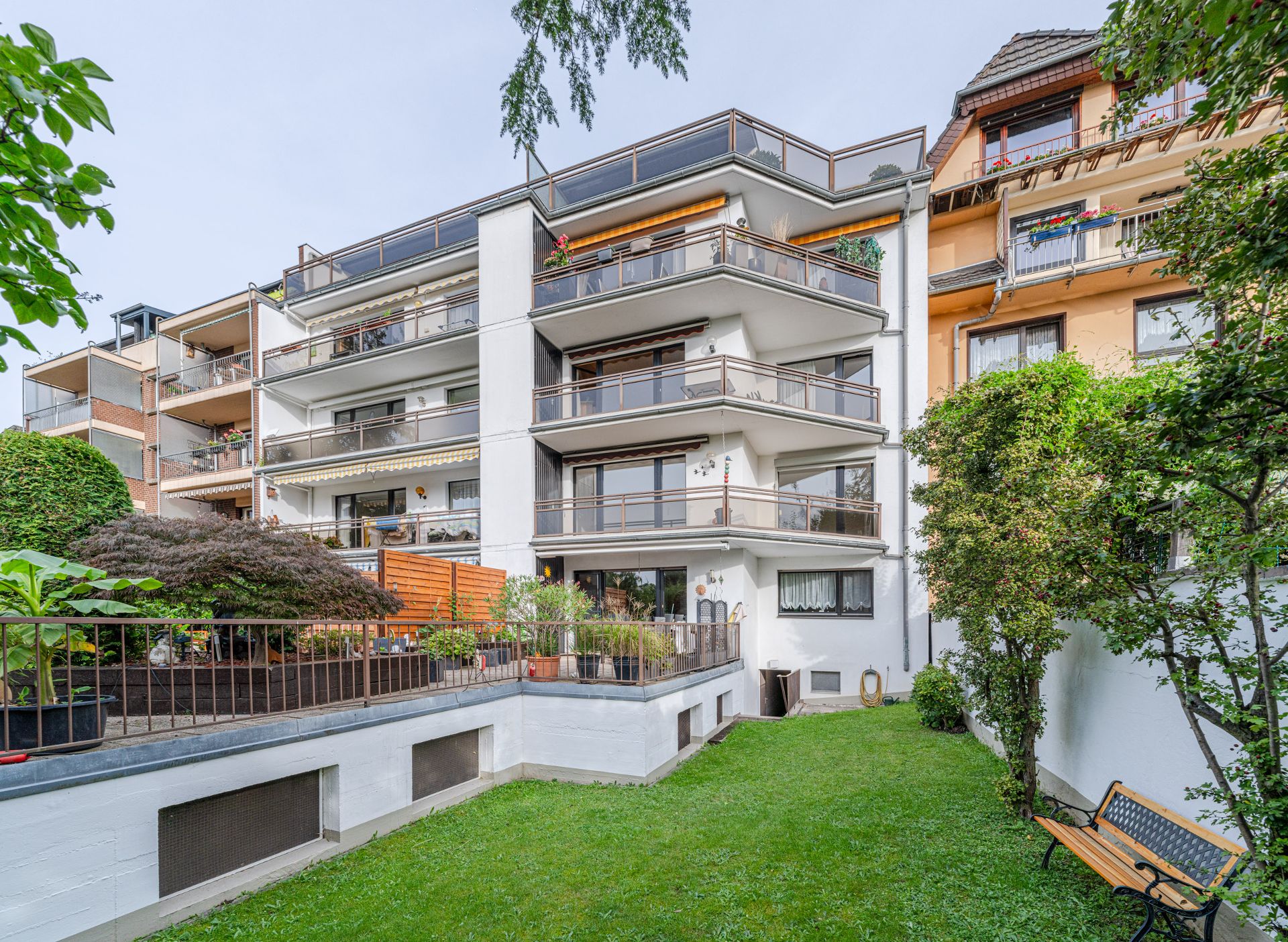Mehrfamilienhaus mit Balkonen und kleinem Gartenbereich mit Bank im Vordergrund.