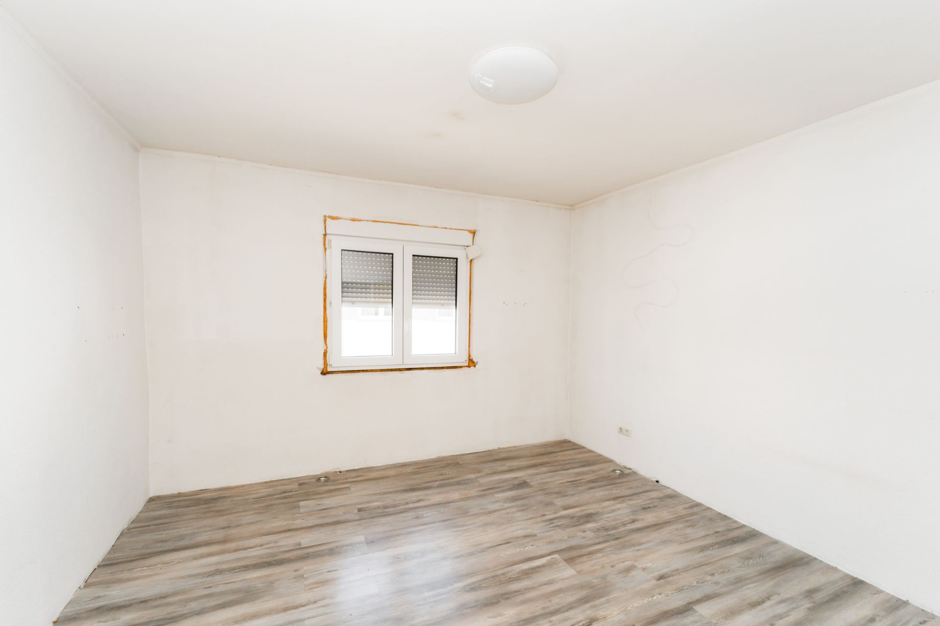 Leerer Raum mit weißen Wänden, Fenster und hellem Laminatboden.