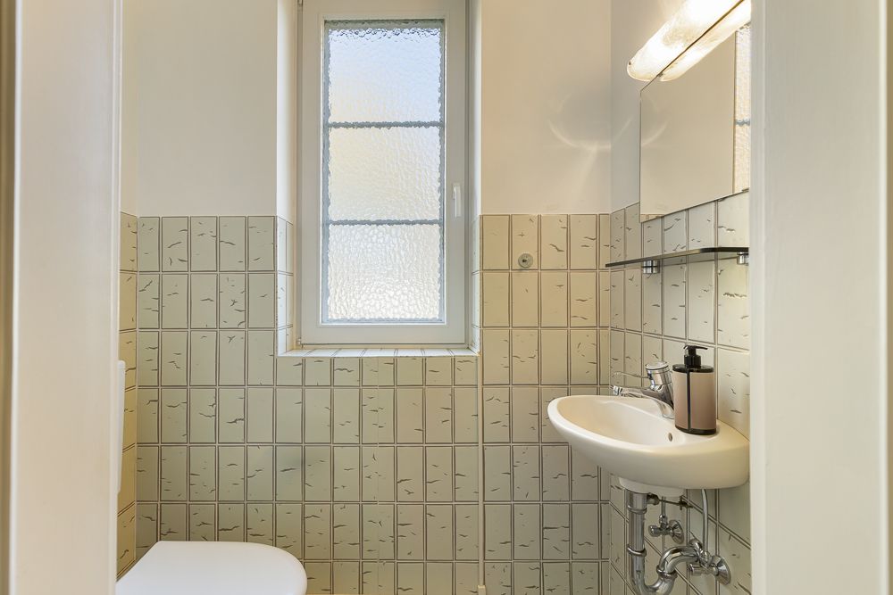 Kleines Badezimmer mit Fenster, Waschbecken, Spiegel und Kachelwänden.
