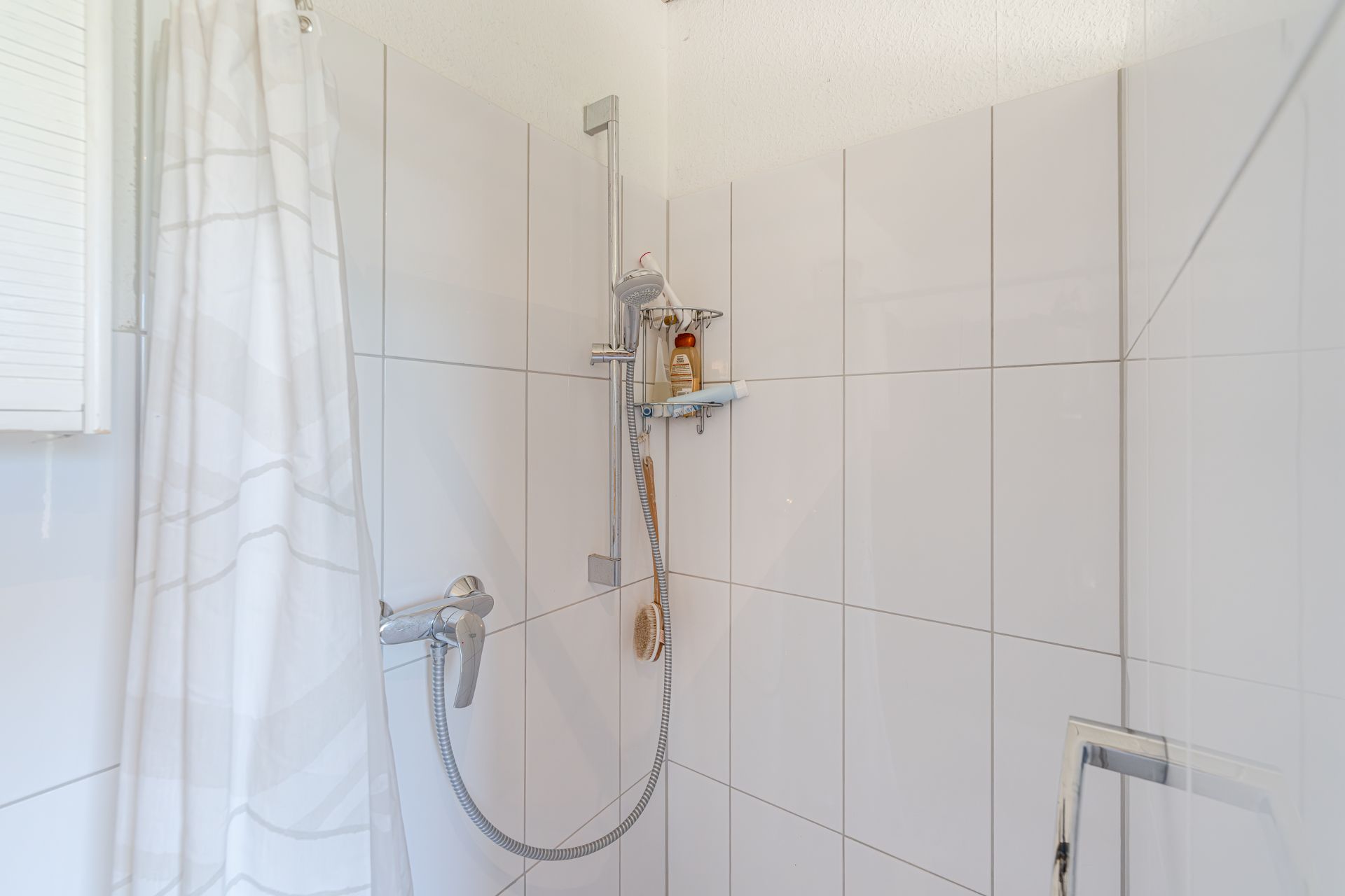 Dusche mit weiß gefliester Wand, Duschkopf, Vorhang und Duschutensilien.