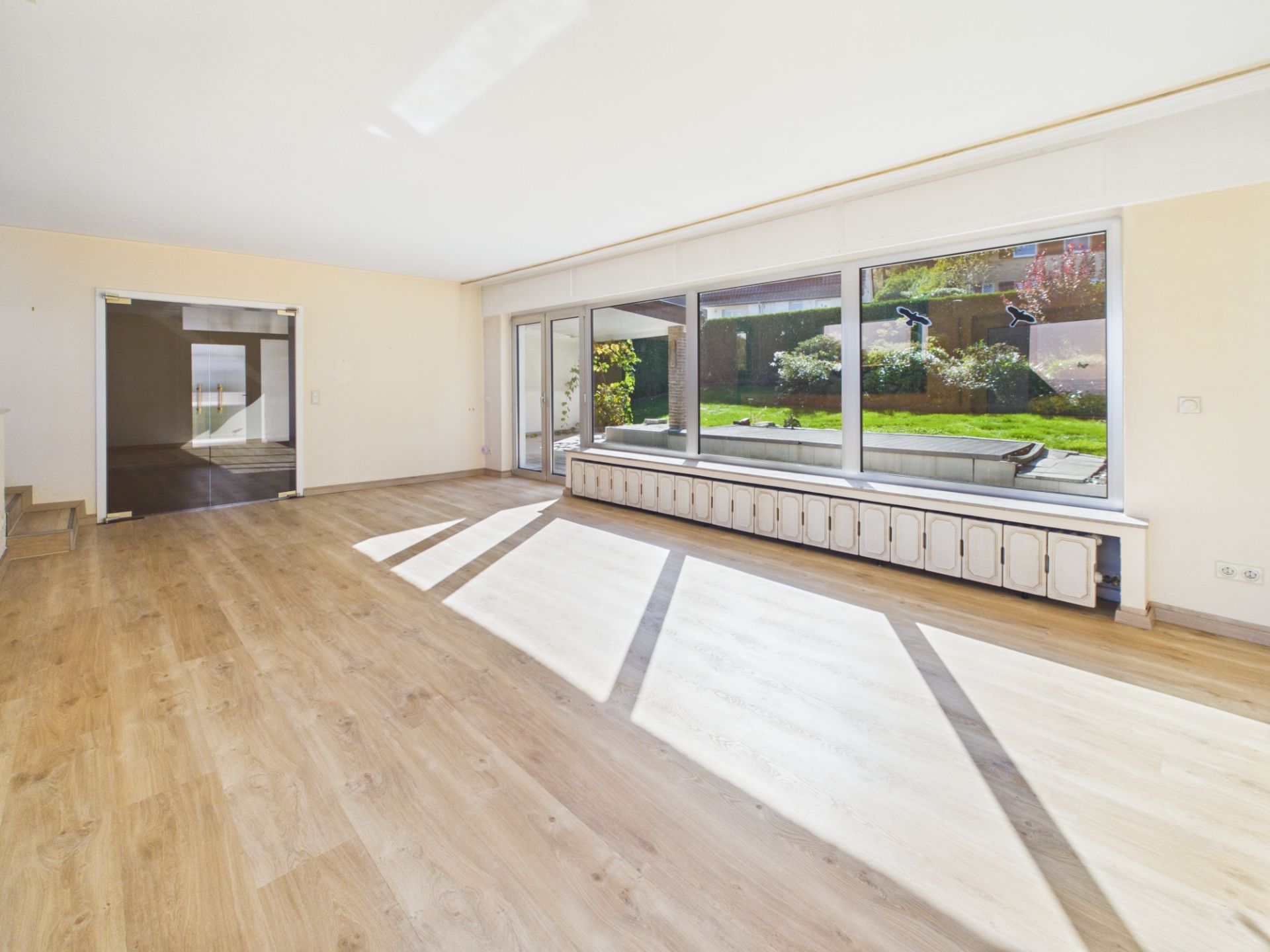 Helles Wohnzimmer mit großen Fenstern, Parkettboden und Blick in einen Garten.