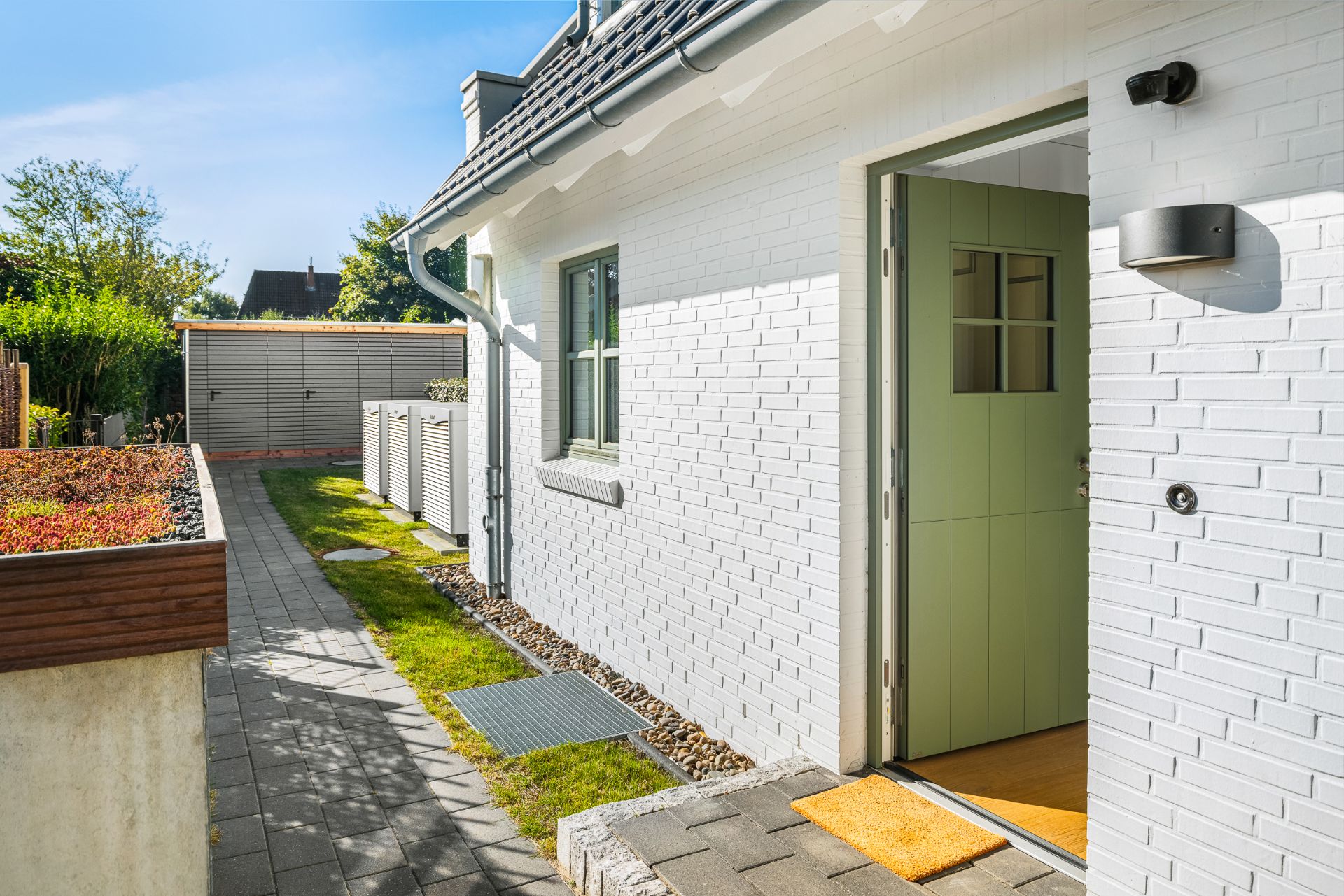 Weiße Hausfassade mit grüner Haustür, teilweise geöffnet, und Weg entlang des Hauses.