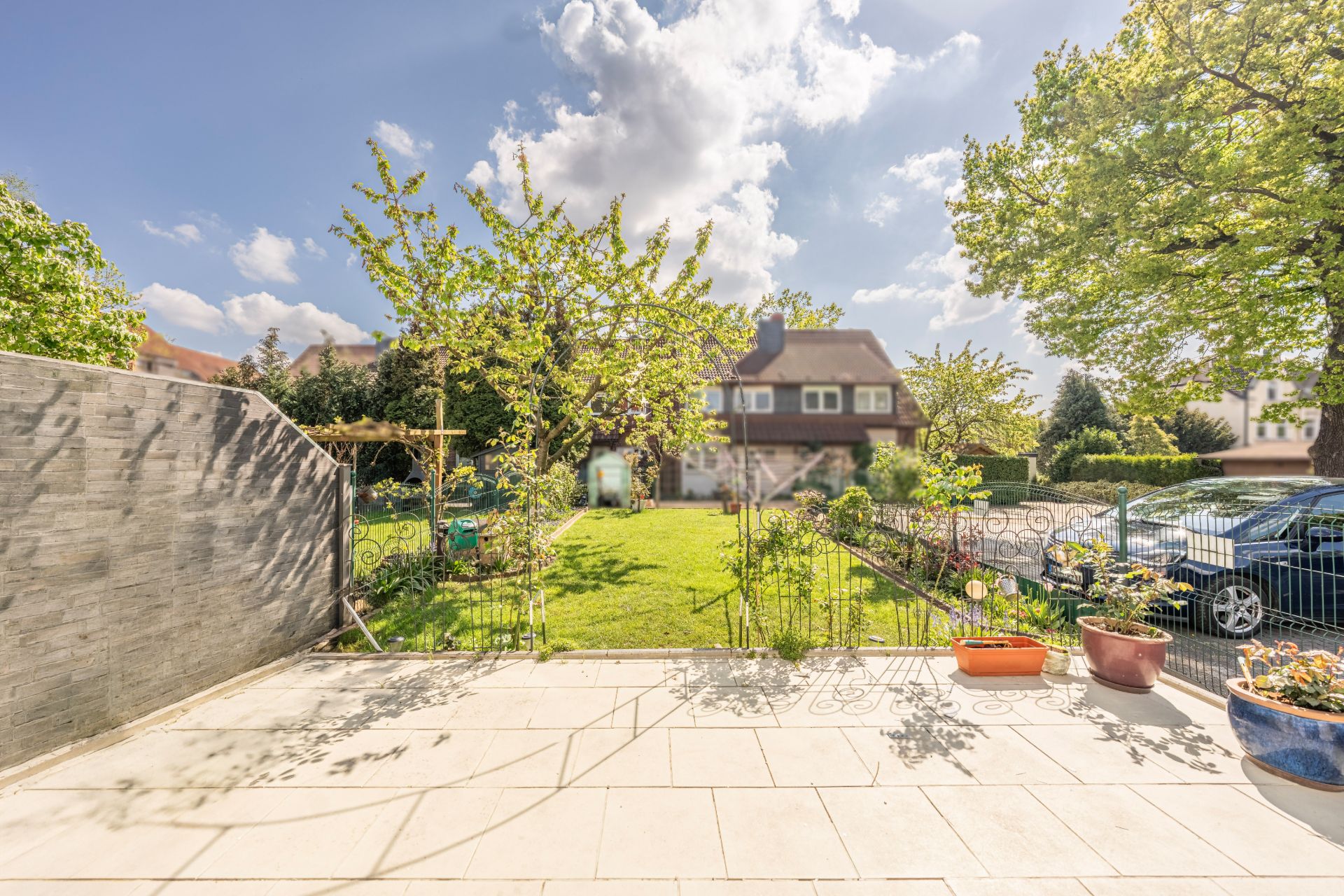 Blick von einer Terrasse auf einen Garten mit Bäumen und Blumen, im Hintergrund ein Wohnhaus.