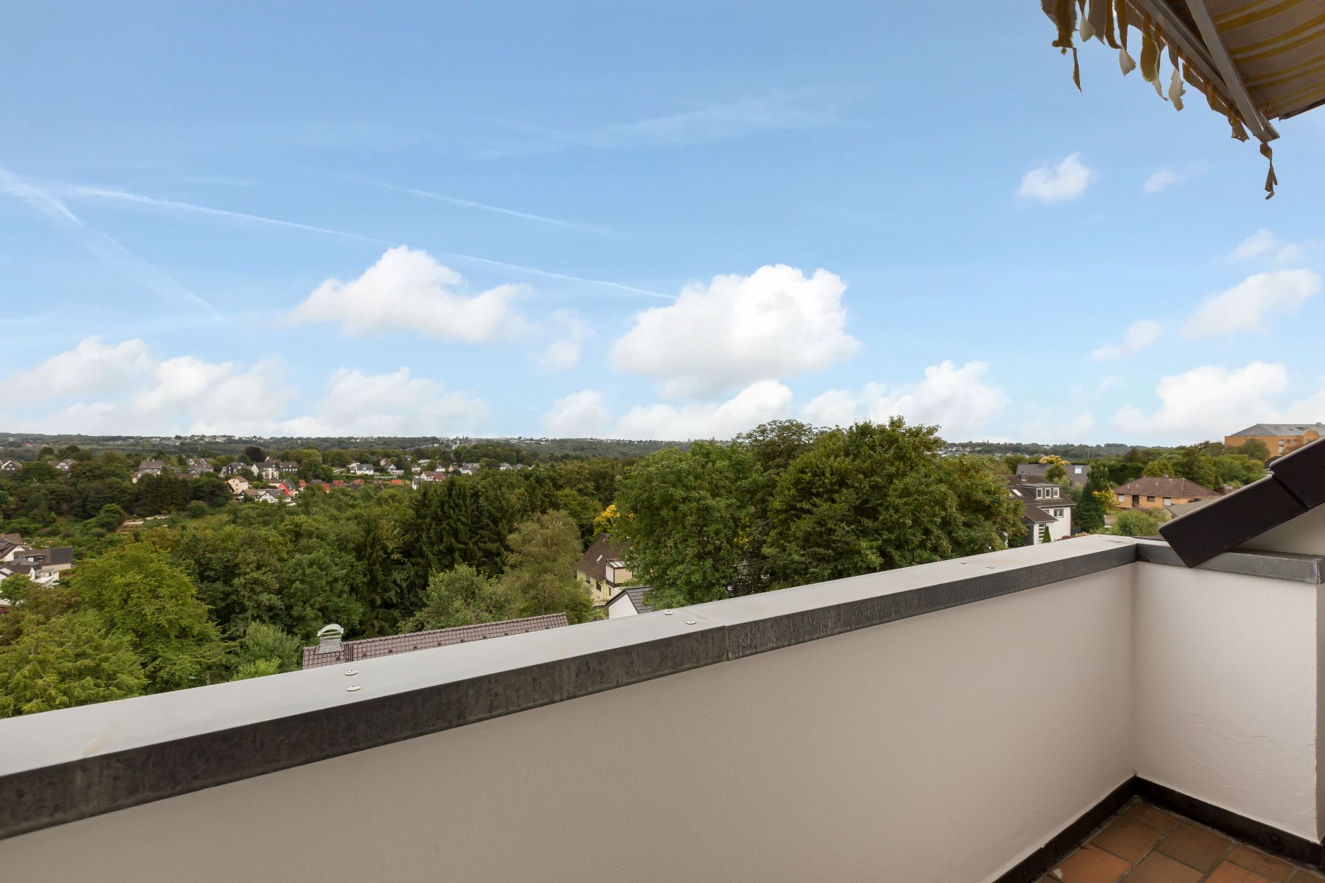 Balkon mit Ausblick auf grüne Bäume und Wohnhäuser unter blauem Himmel.
