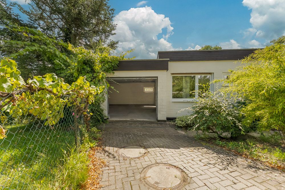 Einfamilienhaus mit Flachdach, Garage und großem Fenster, umgeben von Pflanzen.