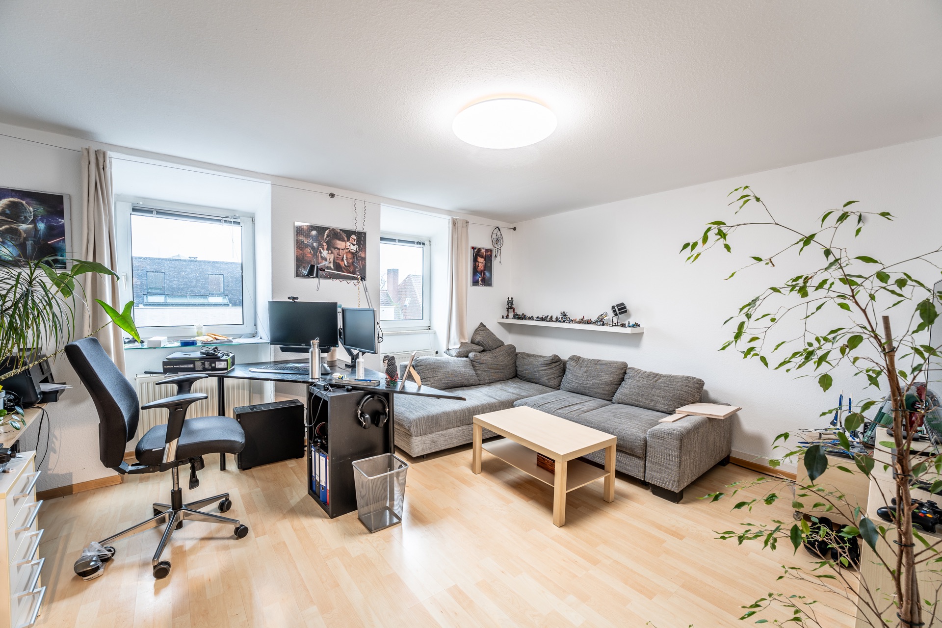 Helles Wohnzimmer mit Schreibtisch, Computern, grauem Sofa, Couchtisch und Pflanze.
