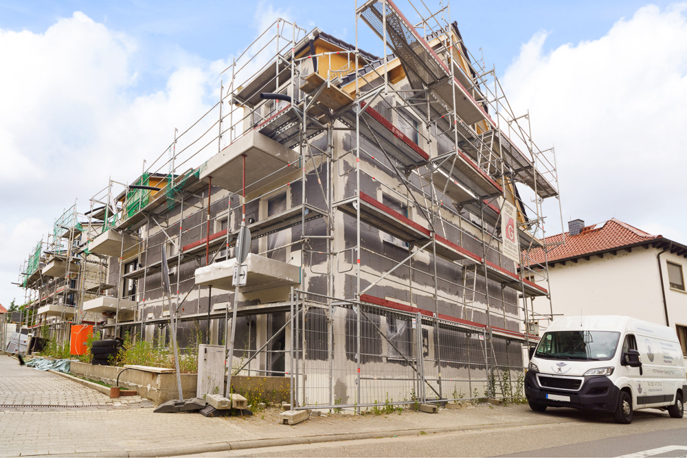 Mehrfamilienhaus im Bau mit Gerüsten an der Fassade, weißer Transporter parkt davor.