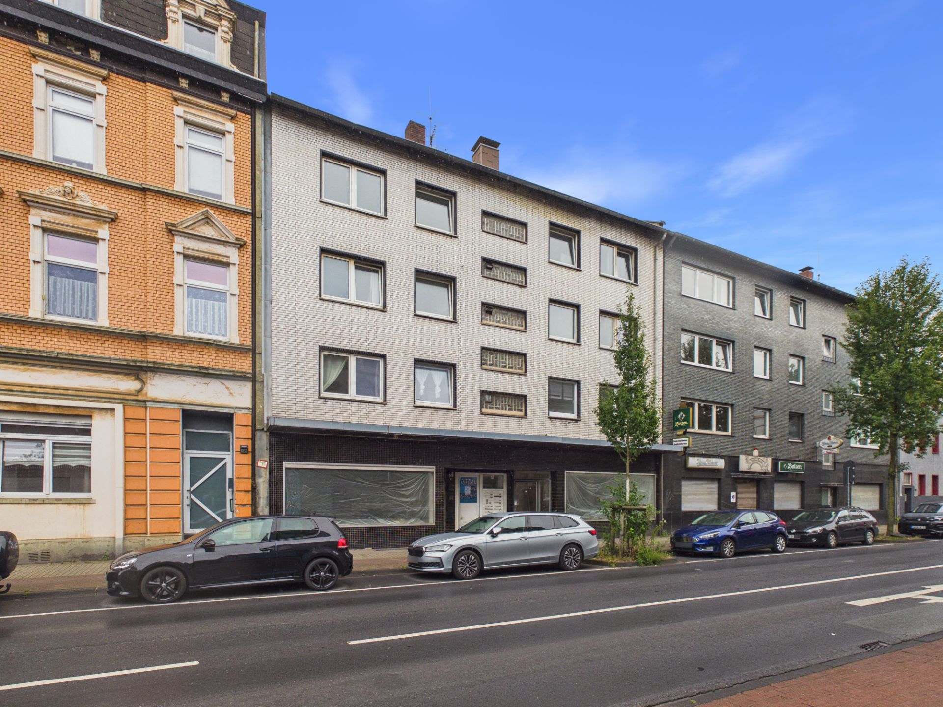 Straßenansicht mehrerer Wohngebäude mit parkenden Autos und Bäumen am Gehweg.