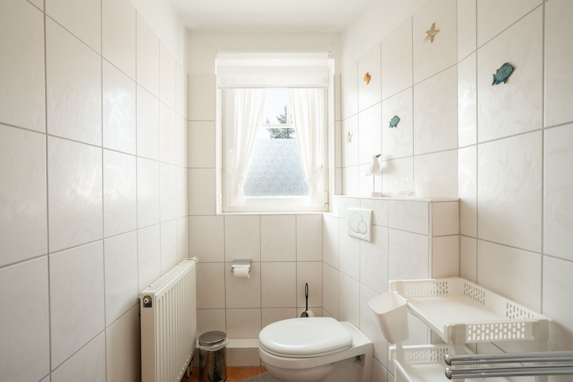 Kleines, weiß gefliestes Bad mit WC, Fenster, Heizkörper und dekorativen Wandstickern.