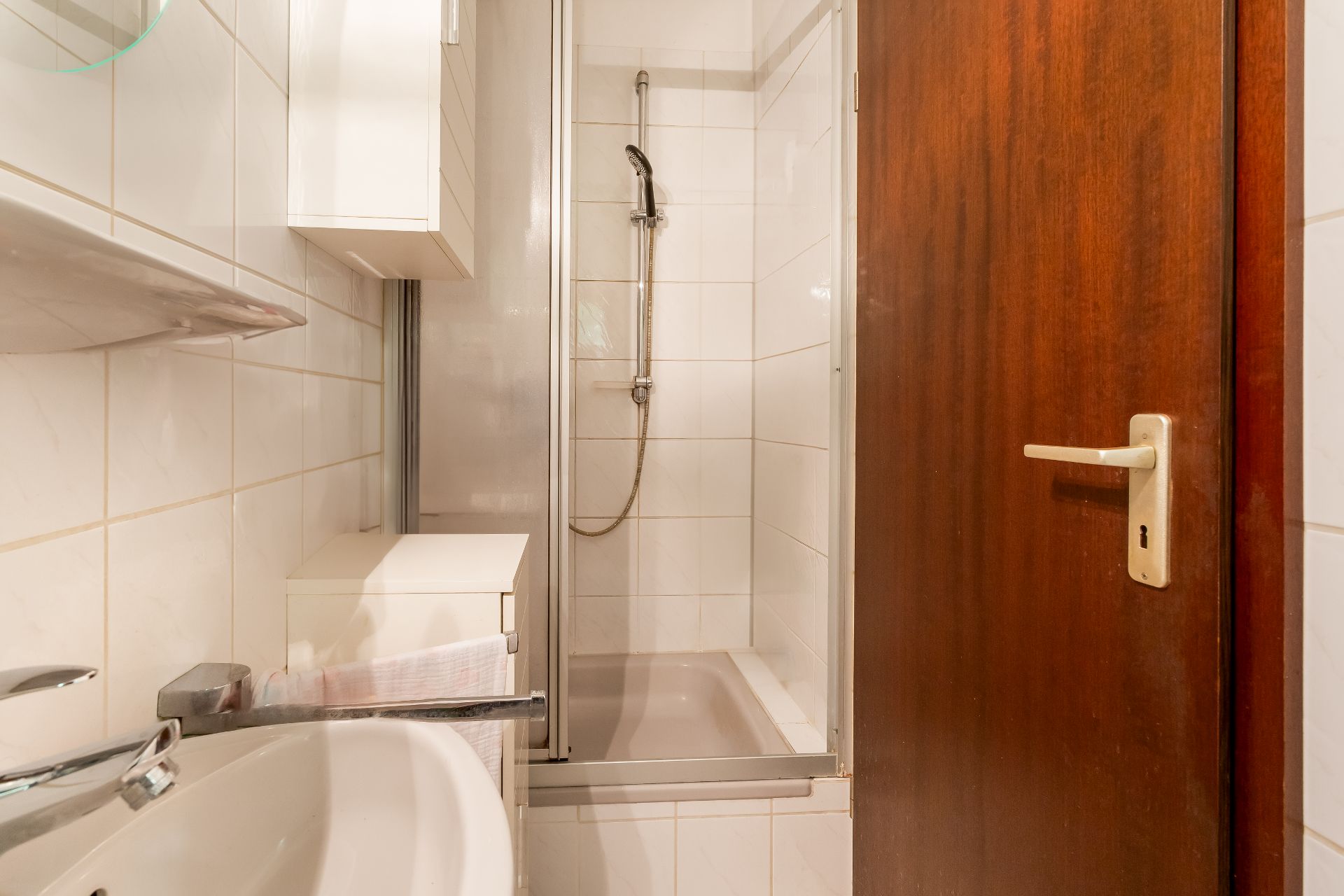 Kleines Bad mit Dusche, Waschbecken und Holzhaustür, weiße Fliesen an den Wänden.