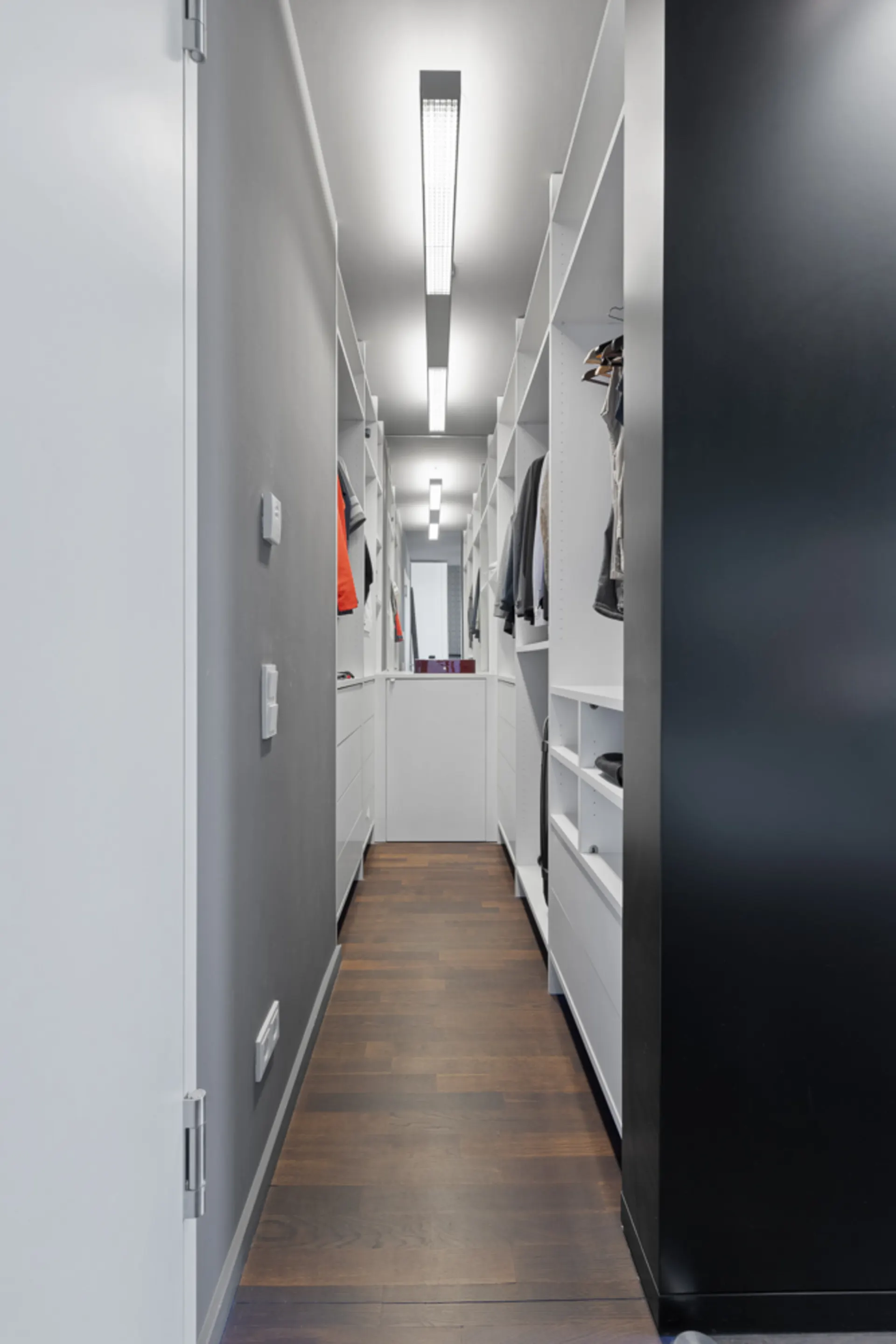 Moderner, schmaler begehbarer Kleiderschrank mit Regalen, Kleidung und Holzfußboden.