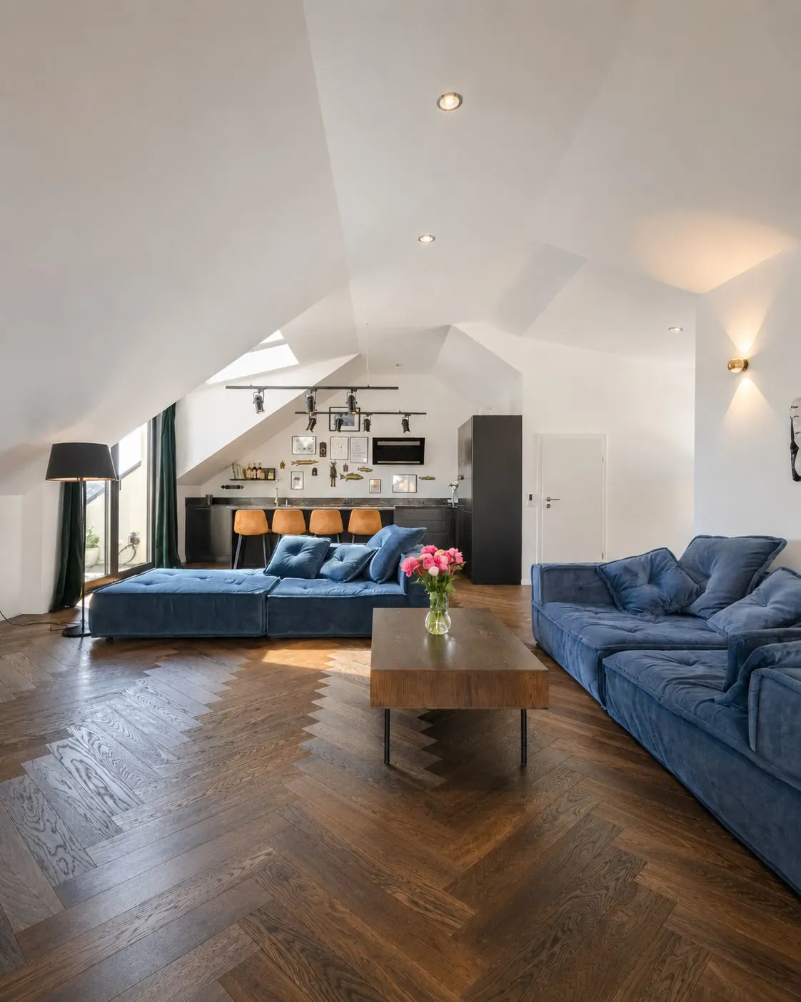 Helles Wohnzimmer mit blauen Sofas, Holztisch und offener Küche im Hintergrund.