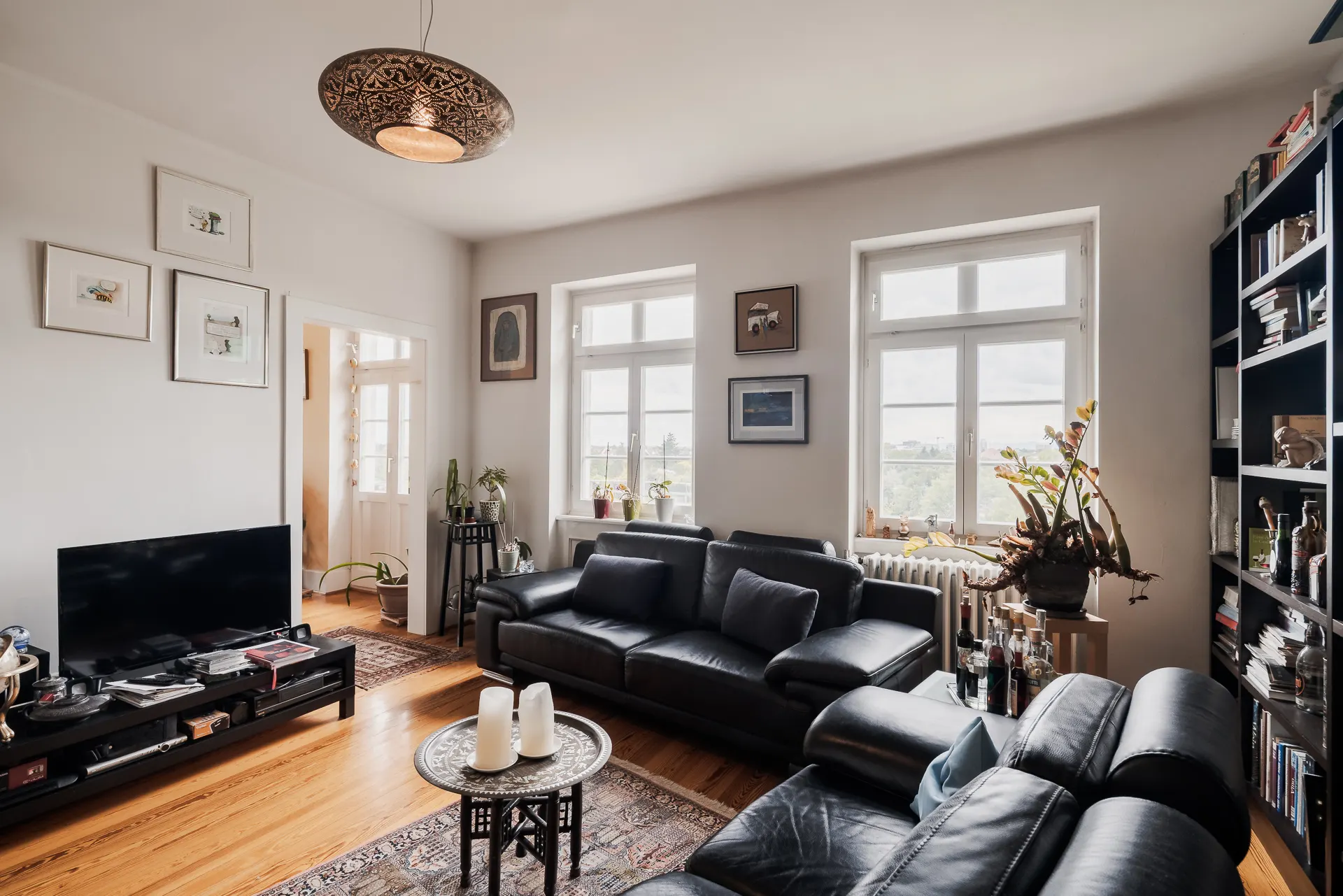 Helles Wohnzimmer mit schwarzen Ledersofas, großen Fenstern, TV und Bücherregal.