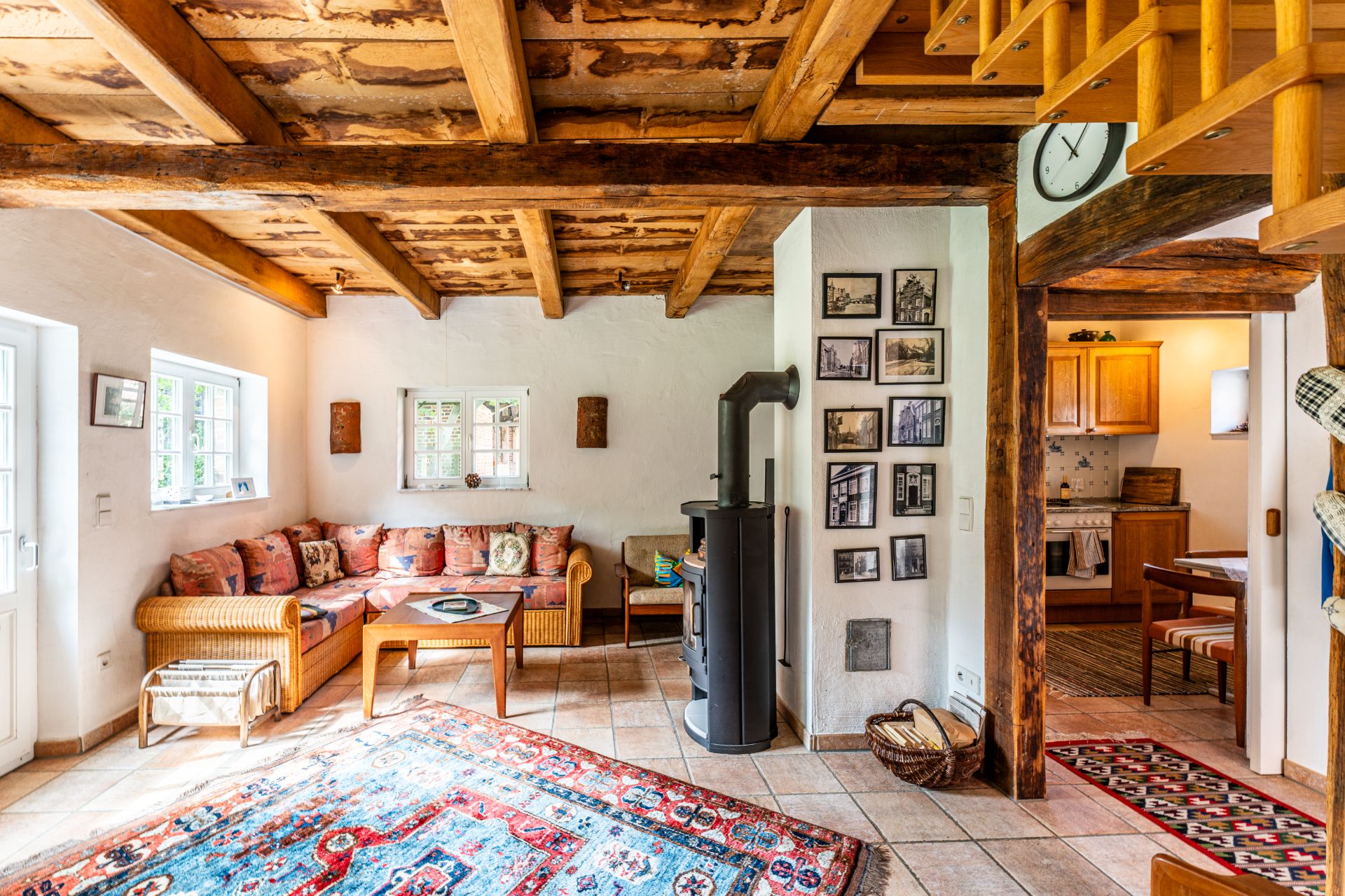 Gemütliches Wohnzimmer mit Holzbalkendecke, Sofa, Kaminofen und Blick in eine offene Küche.