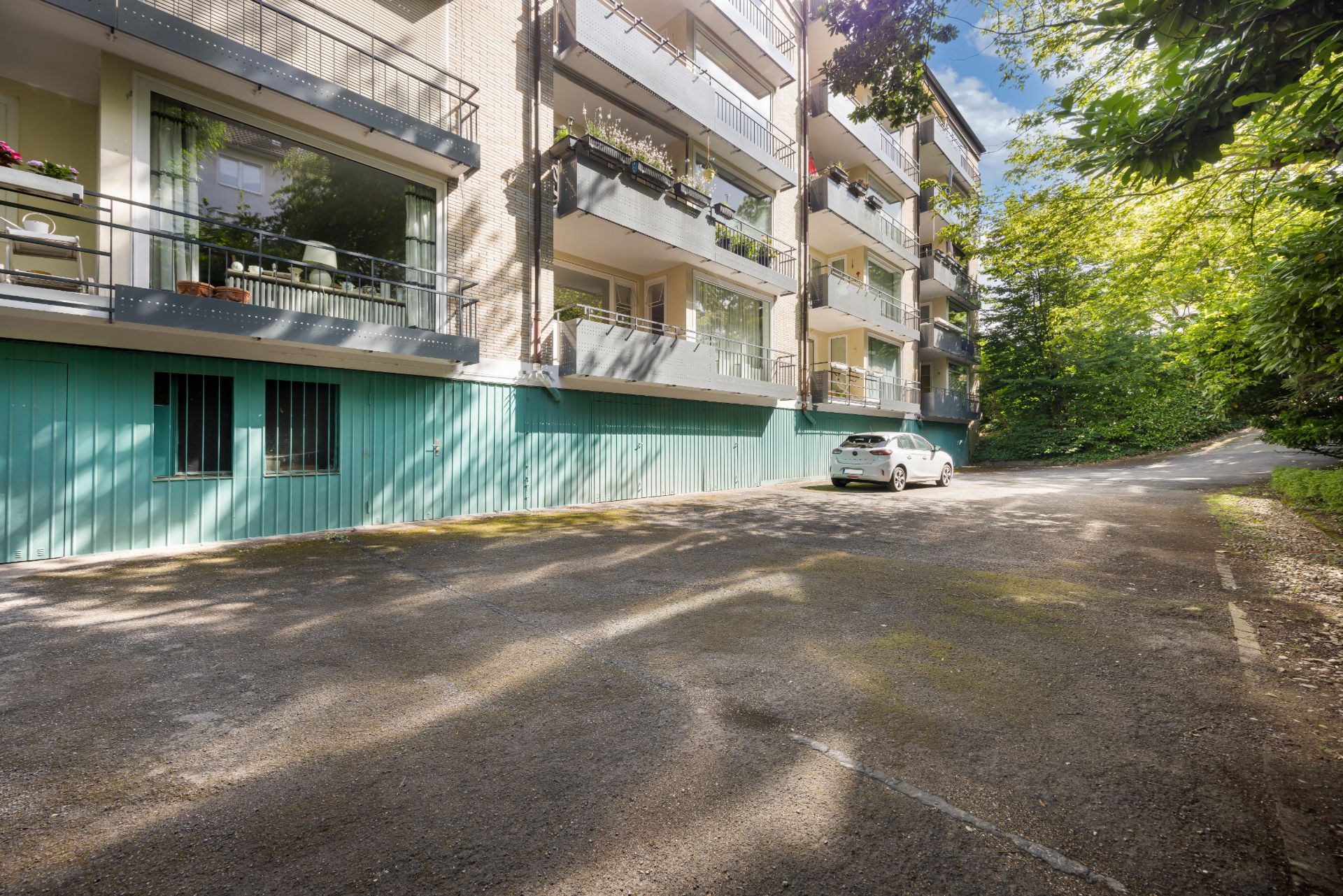 Mehrfamilienhaus mit Balkonen und Parkplatz, ein Auto steht auf dem Parkplatz, umgeben von Bäumen.