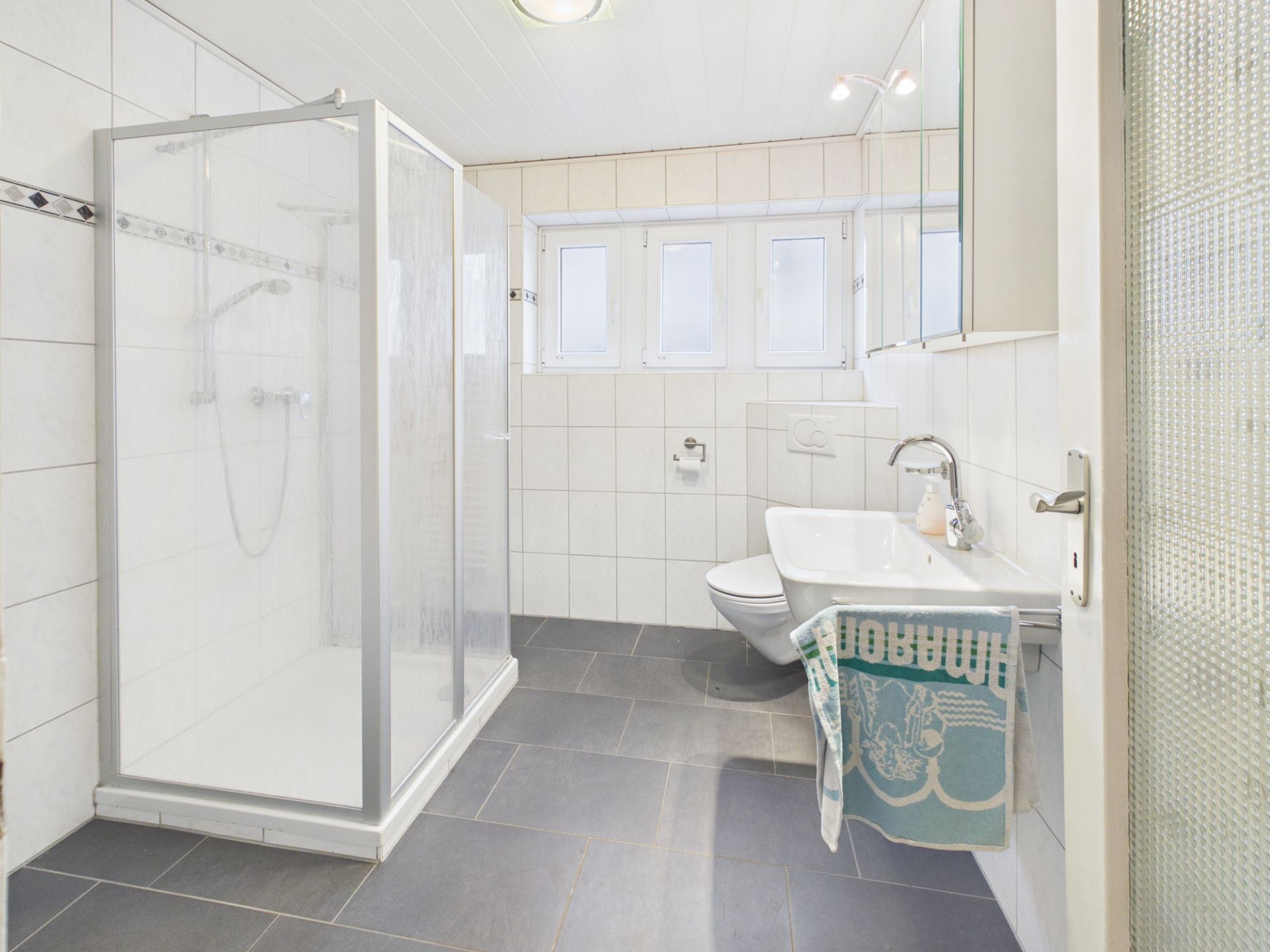 Helles Badezimmer mit Dusche, WC und Waschbecken, grauen Fliesen und Handtuch am Waschbecken.