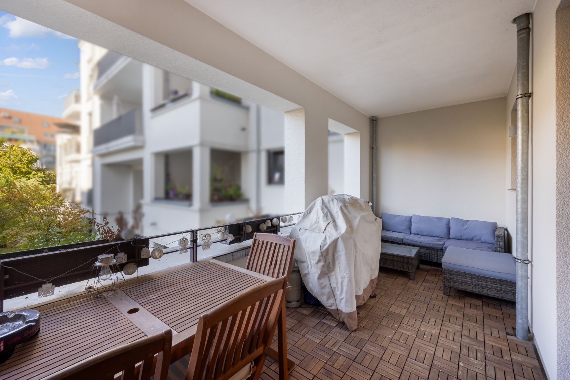 Balkon mit Holztisch, Stühlen, grauer Eckcouch und Grill unter Abdeckung.