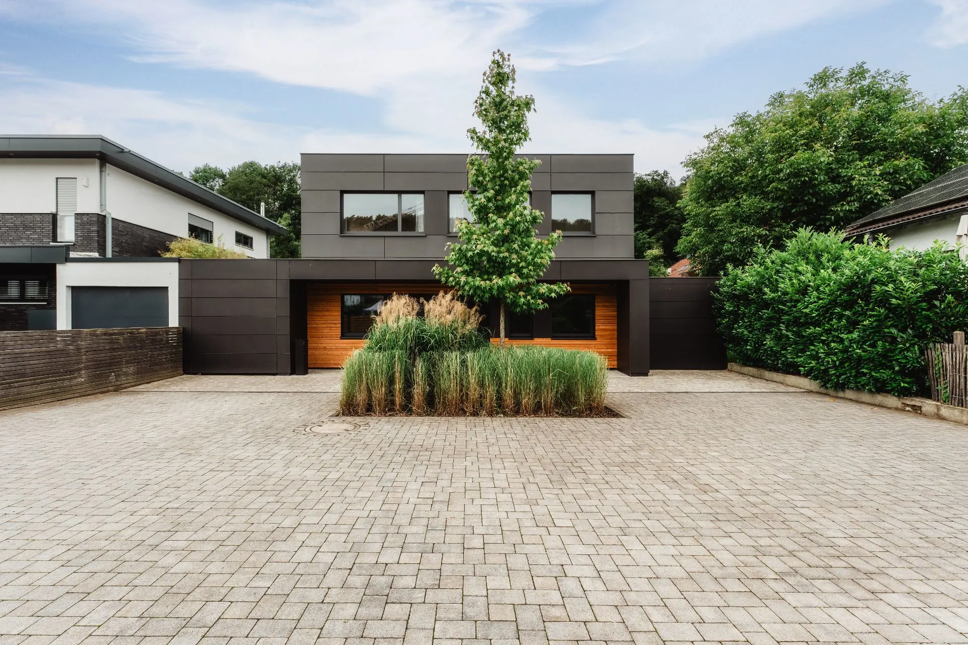 Modernes Wohnhaus mit dunkler Fassade, großen Fenstern und Baum im Vorgarten.