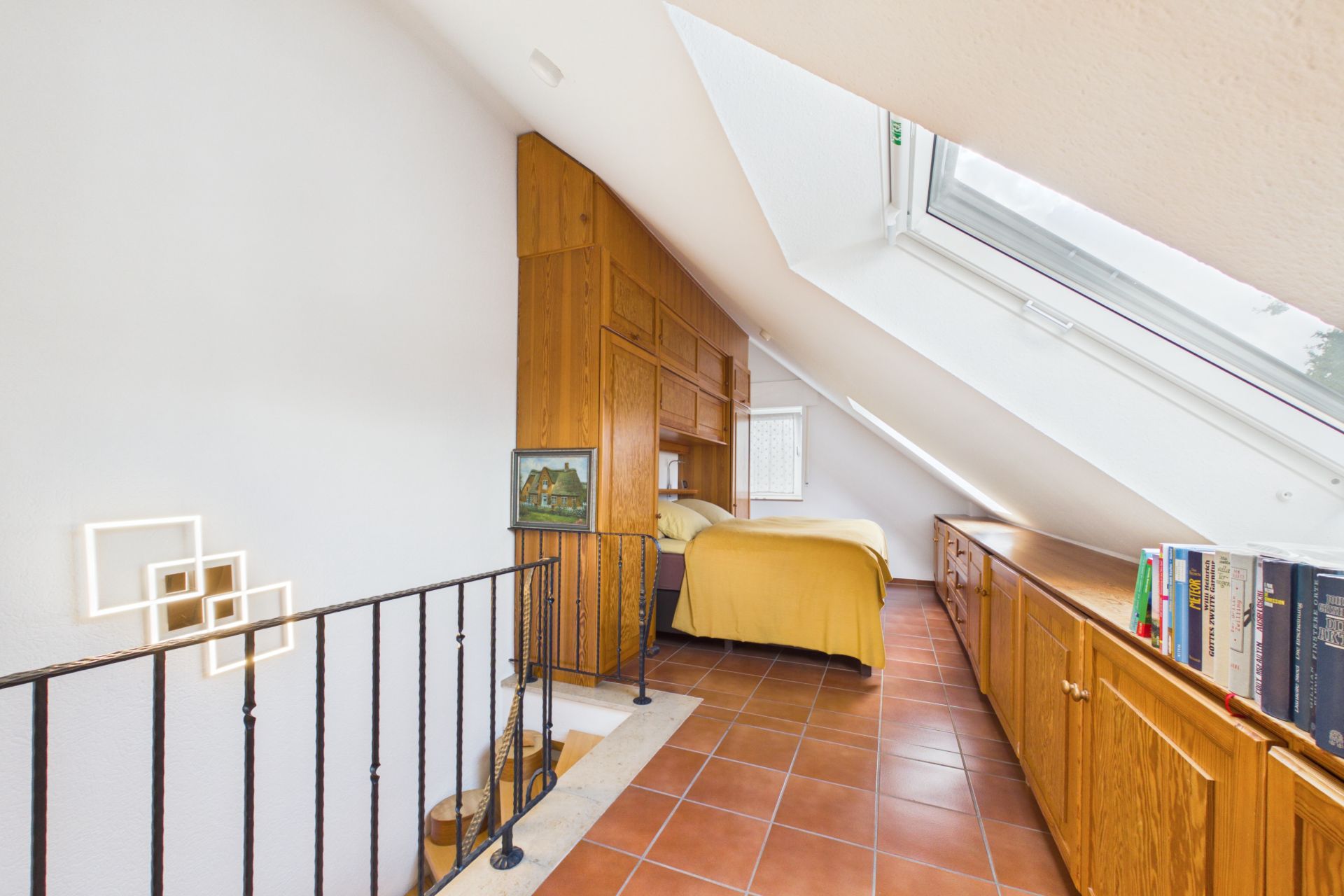 Helles Dachzimmer mit Bett, Holzschränken, Bücherregal und großem Dachfenster.