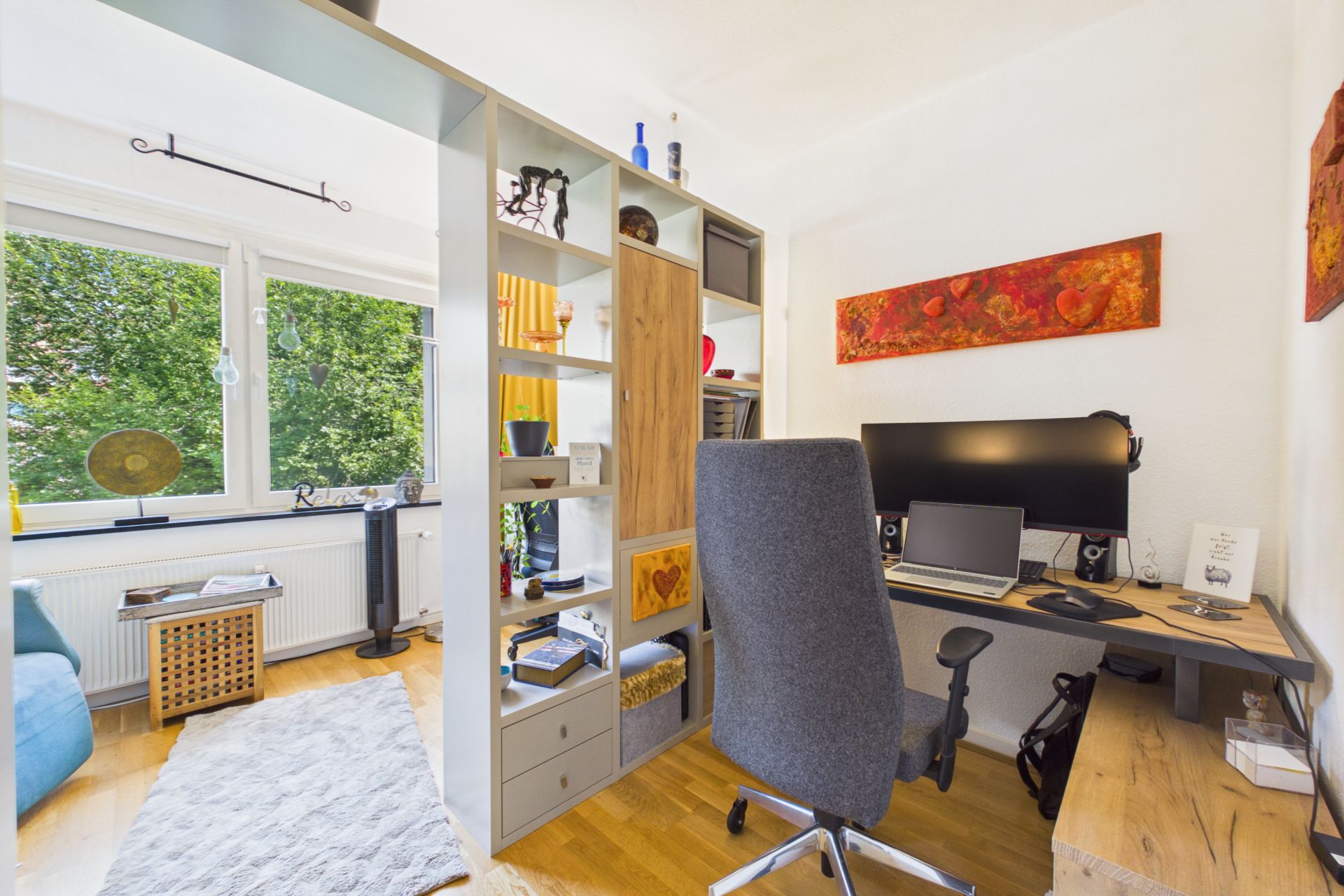 Helles Arbeitszimmer mit Schreibtisch, Regal und großem Fenster, das auf grüne Bäume blickt.