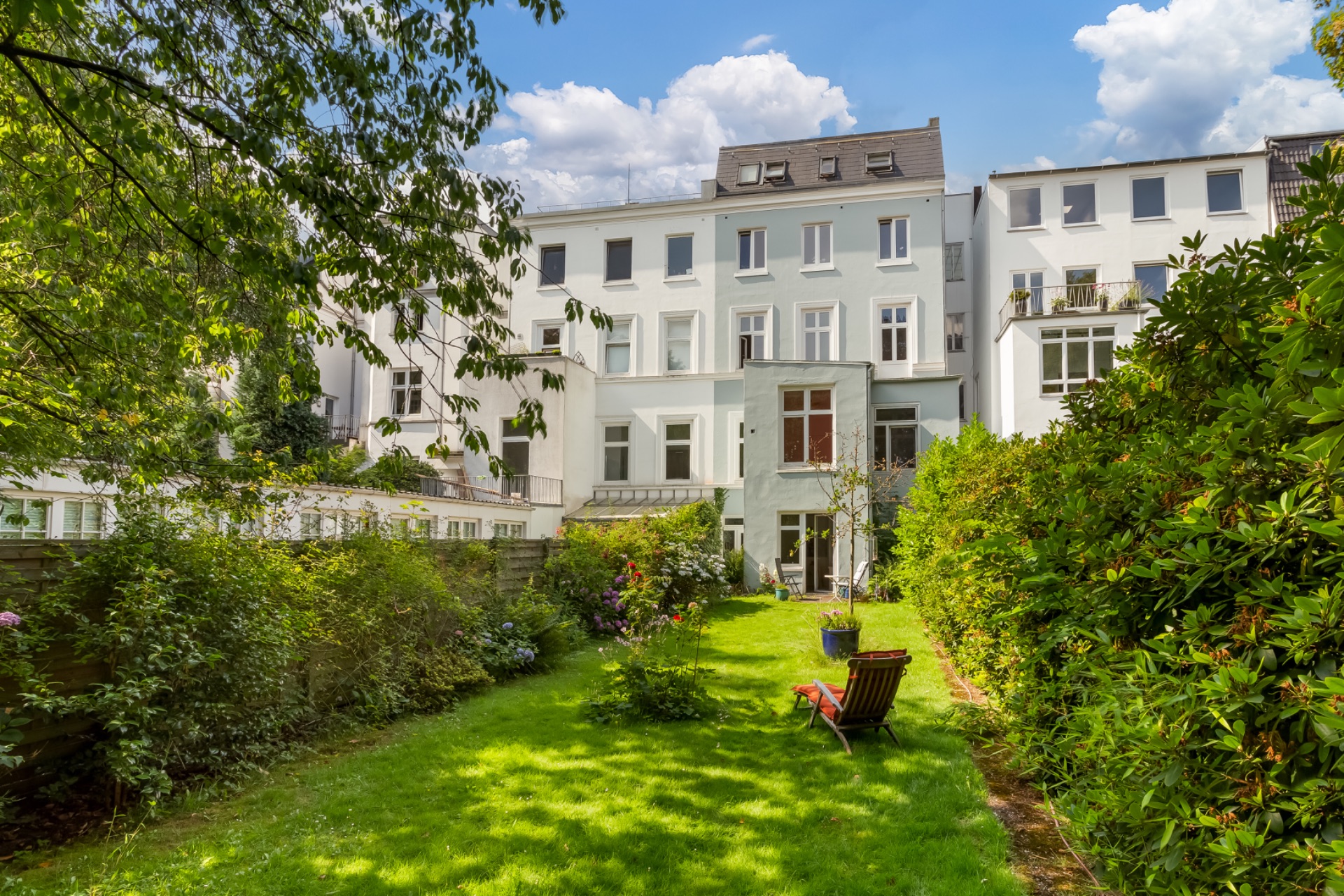 Grüner Garten mit Liegestuhl hinter einem mehrstöckigen Wohnhaus bei Tageslicht.