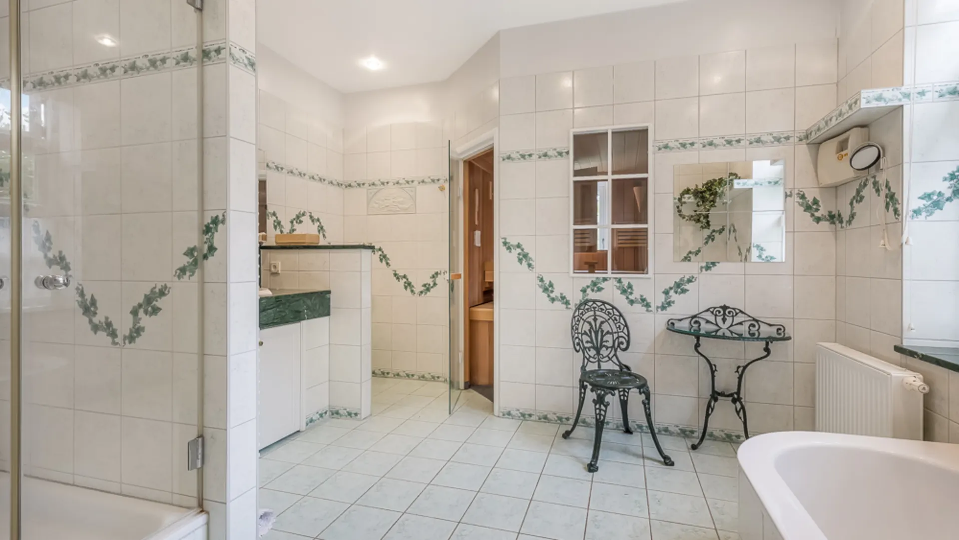 Gefliestes Badezimmer mit Dusche, Badewanne, Stuhl, Tisch und Spiegel. Wände mit grünem Muster verziert.