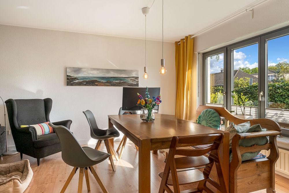 Heller Essbereich mit Holztisch, Stühlen, Sessel, gelben Vorhängen und großen Fenstern.