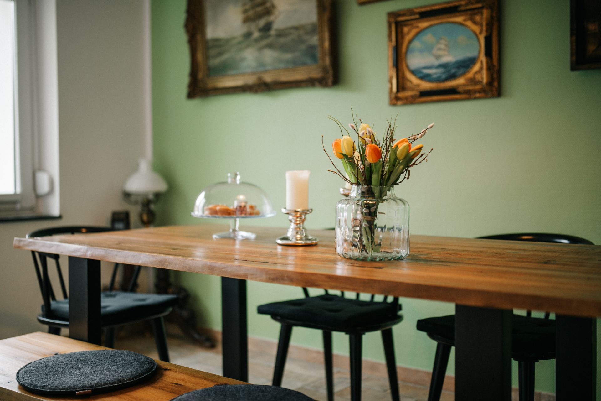 Holztisch mit Vase voller Tulpen und Kerze, Stühle und Gemälde an der grünen Wand.