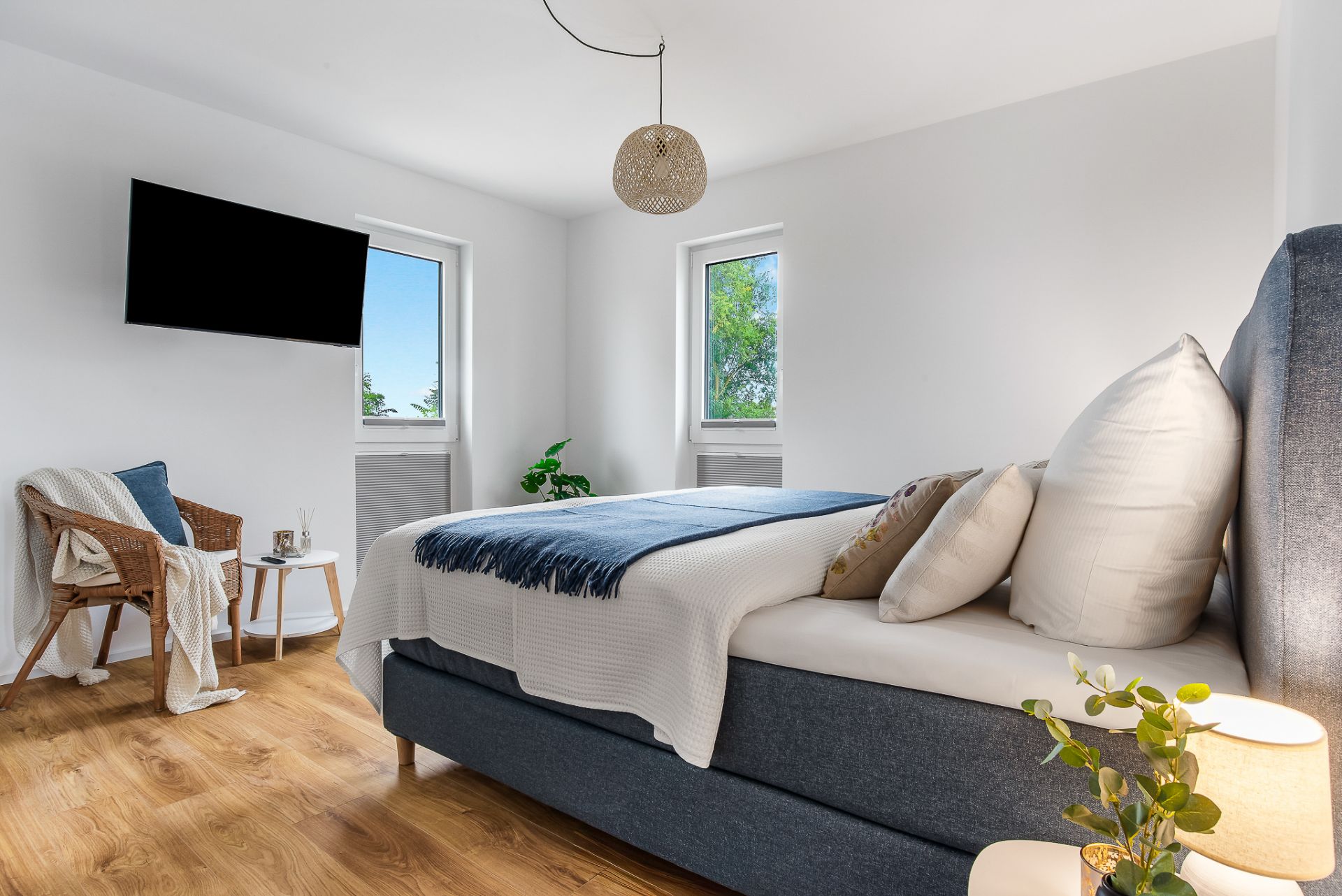 Helles Schlafzimmer mit großem Bett, Holzfußboden, zwei Fenstern und einem Sessel neben einem kleinen Tisch.