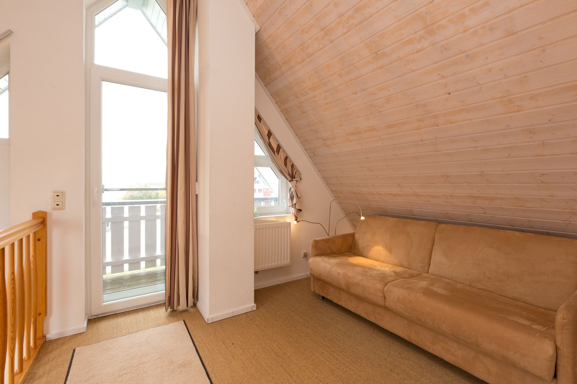 Wohnbereich mit beiger Couch unter schräger Holzdecke, Fenster und Tür zum Balkon.