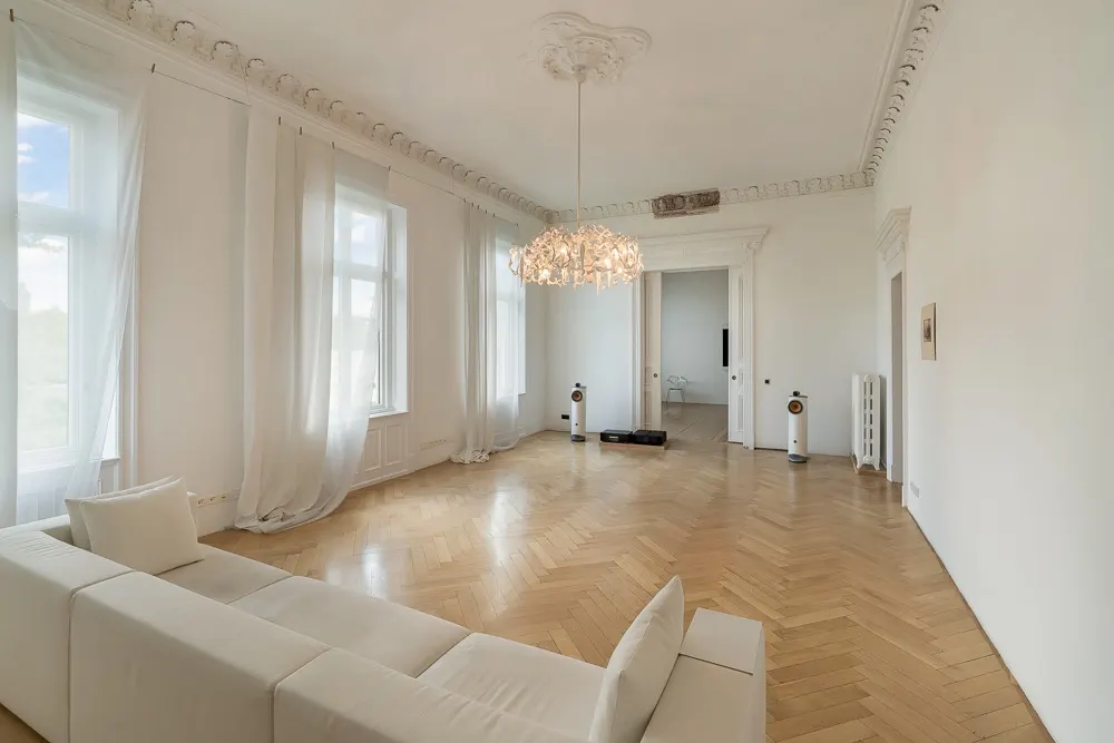 Helles Wohnzimmer mit Parkettboden, weißer Couch, großen Fenstern und Kronleuchter.