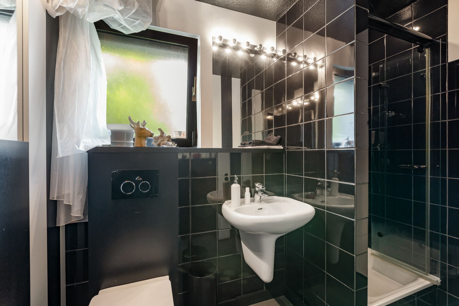 Modernes Badezimmer mit schwarzer Fliesenwand, Waschbecken, WC und Dusche, Spiegel mit Lichtern.