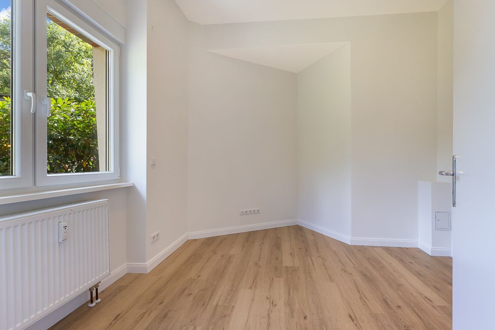 Helles, leeres Zimmer mit Holzfußboden, weißer Wand und großem Fenster.