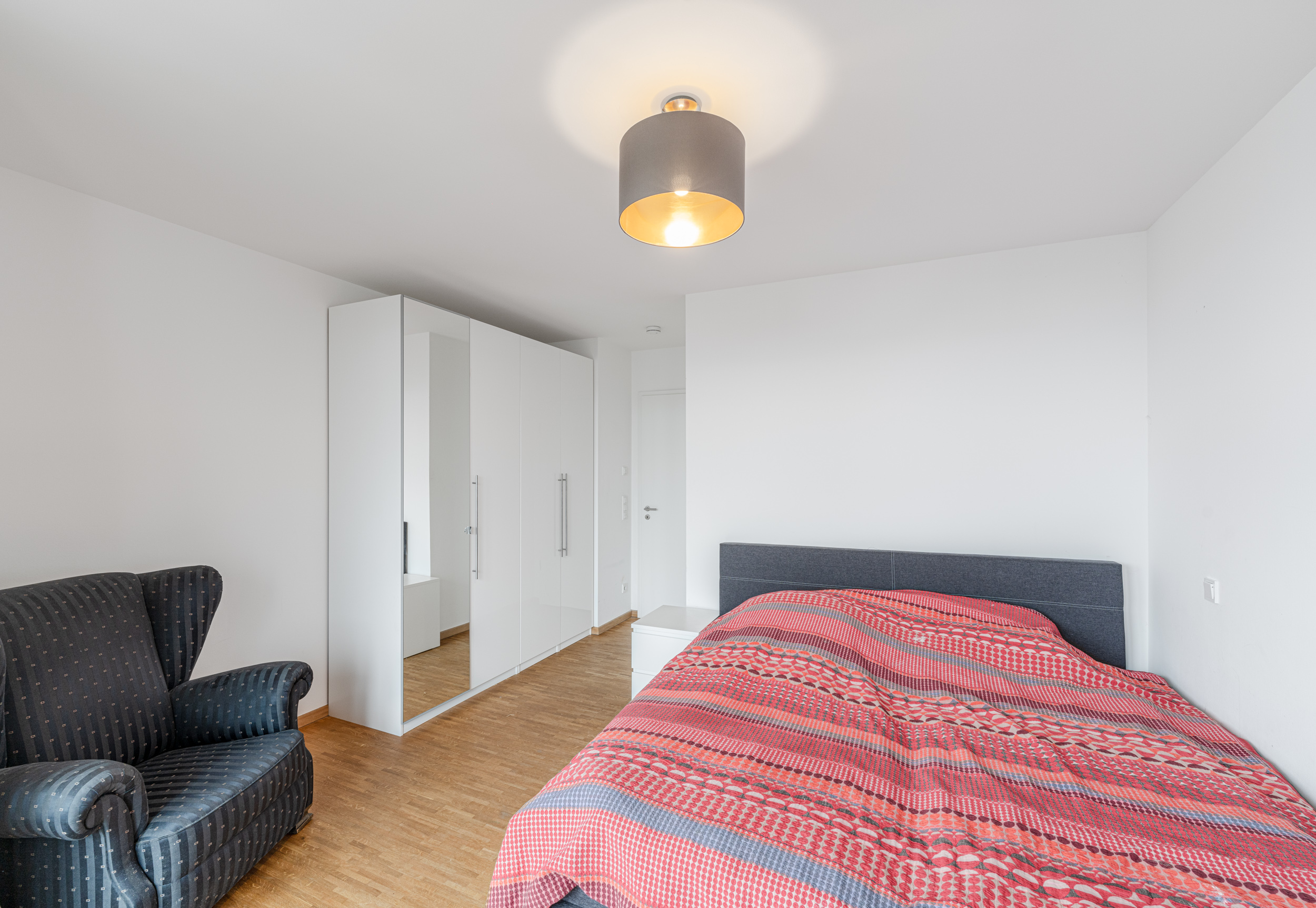 Modernes Schlafzimmer mit rotem Bett, weißem Kleiderschrank und dunklem Sessel.