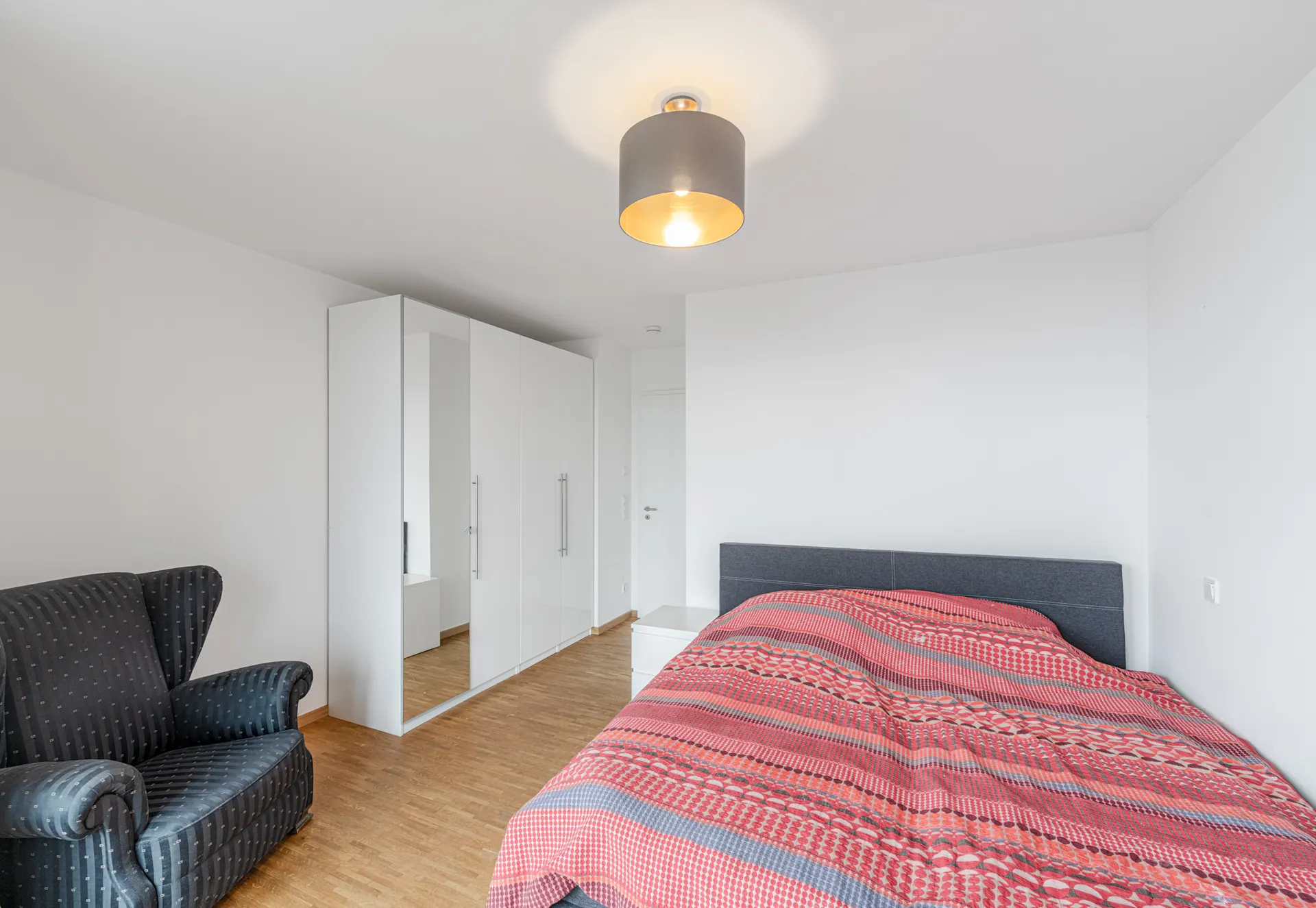 Modernes Schlafzimmer mit rotem Bett, weißem Kleiderschrank und dunklem Sessel.