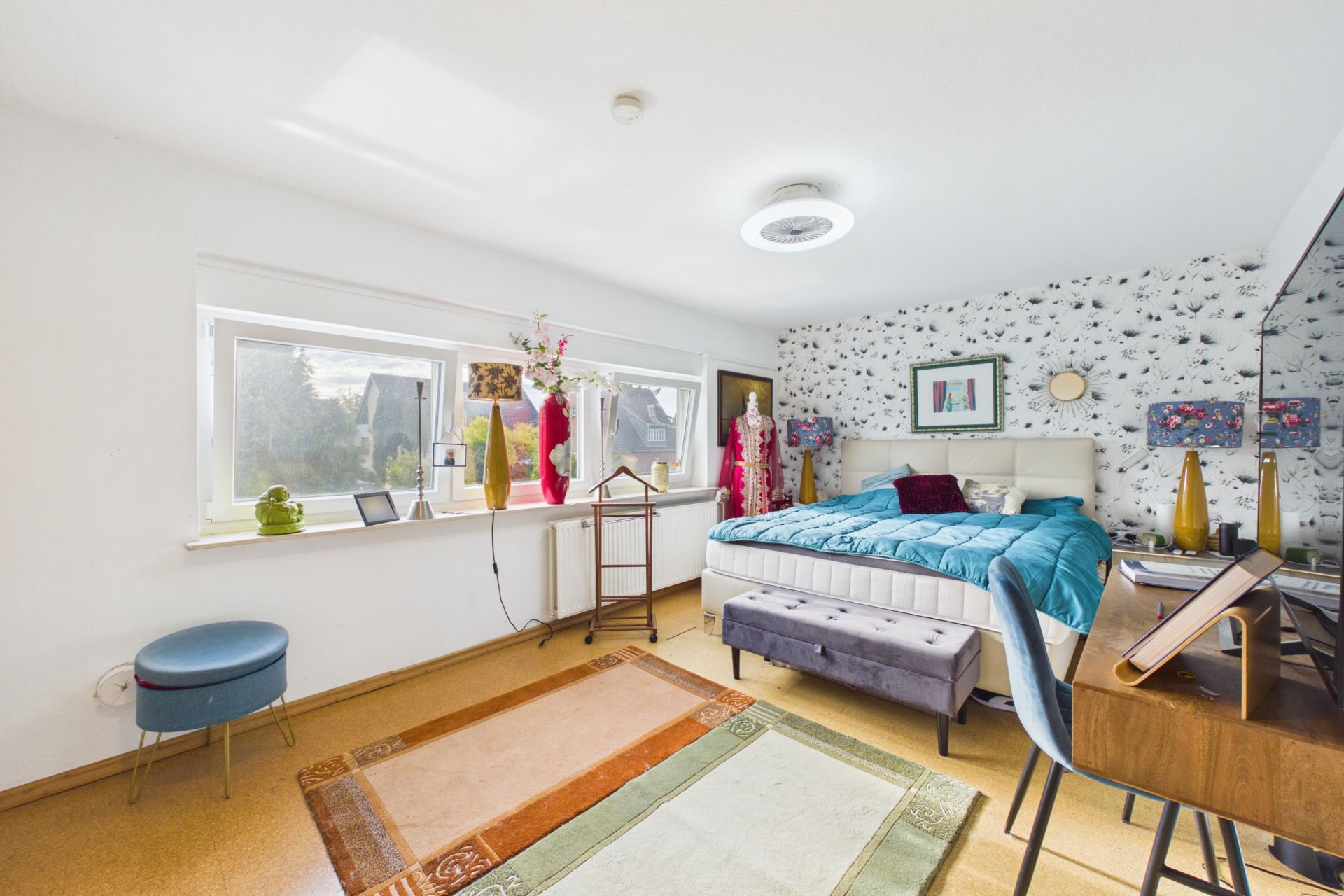Helles Schlafzimmer mit großem Bett, Schreibtisch, Teppich und dekorativen Vasen am Fenster.