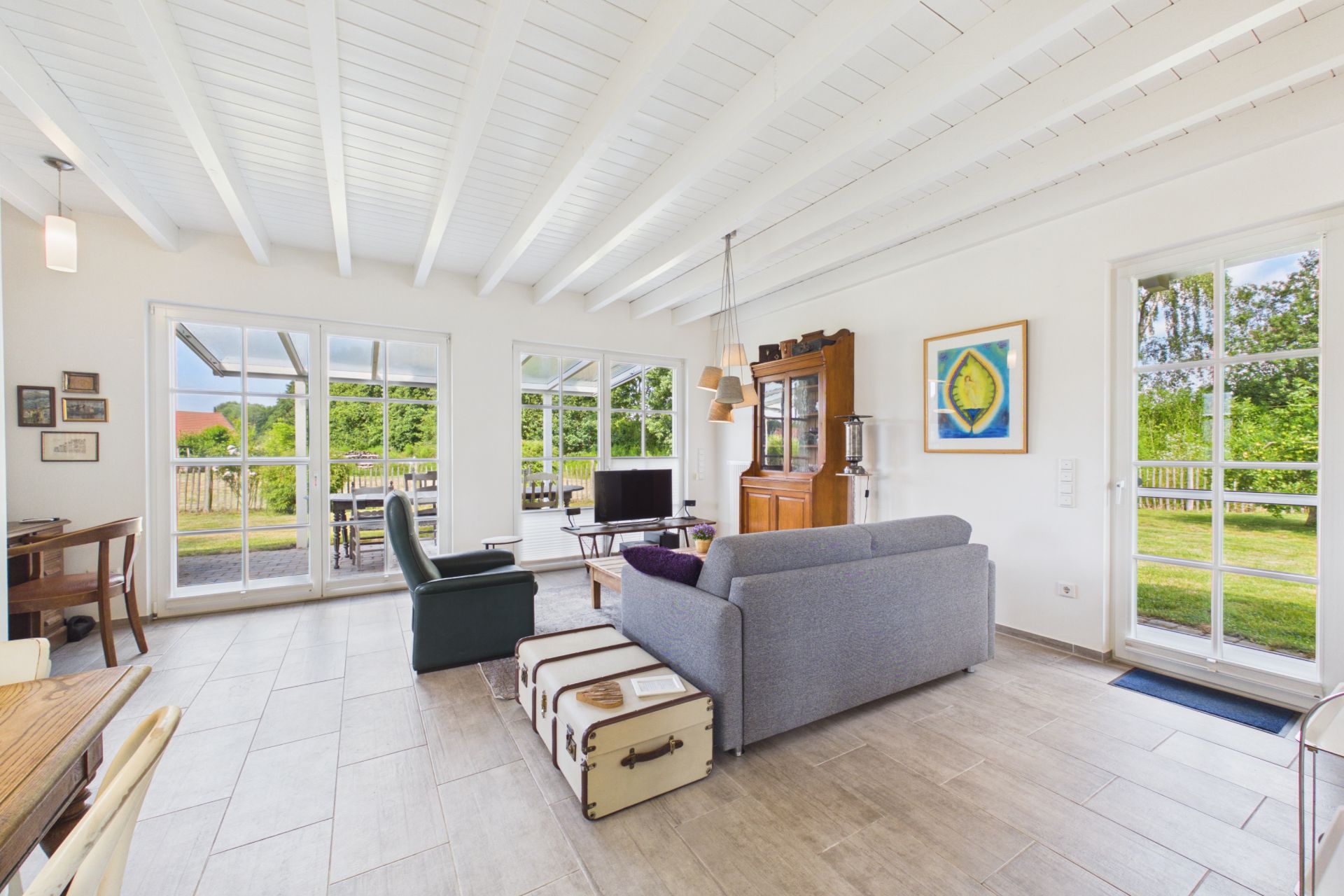 Helles Wohnzimmer mit Sofas, Sessel, Fernseher, großen Fenstern und Blick auf Garten.