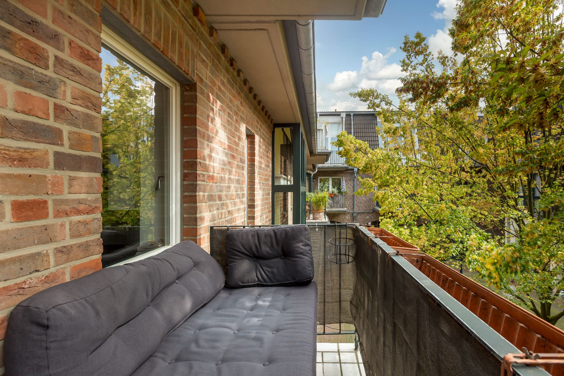 Balkon mit einer grauen Couch, Backsteinwand und Ausblick auf Bäume und Nachbarbalkone.
