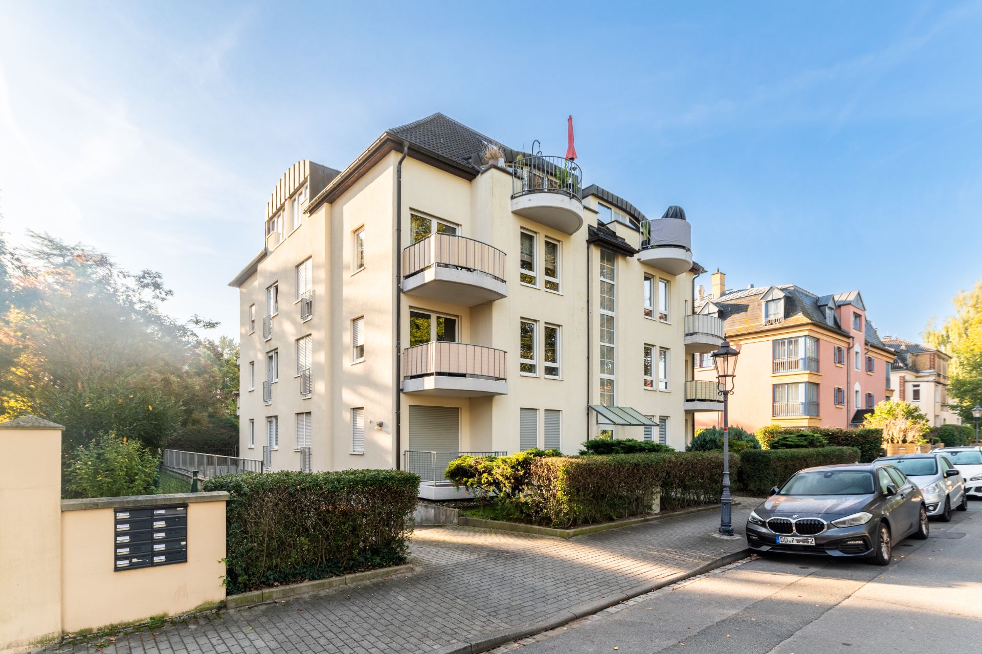 Modernes, helles Wohnhaus mit mehreren Balkonen an einer ruhigen Straße, Autos sind am Straßenrand geparkt.