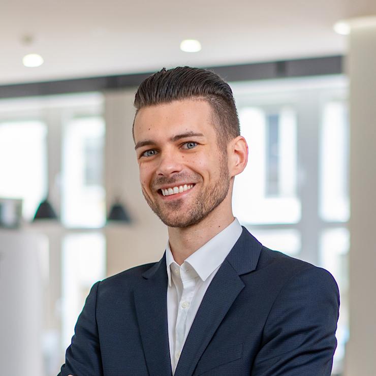 Ein Mann im Anzug steht lächelnd mit verschränkten Armen in einem modernen, hellen Raum.