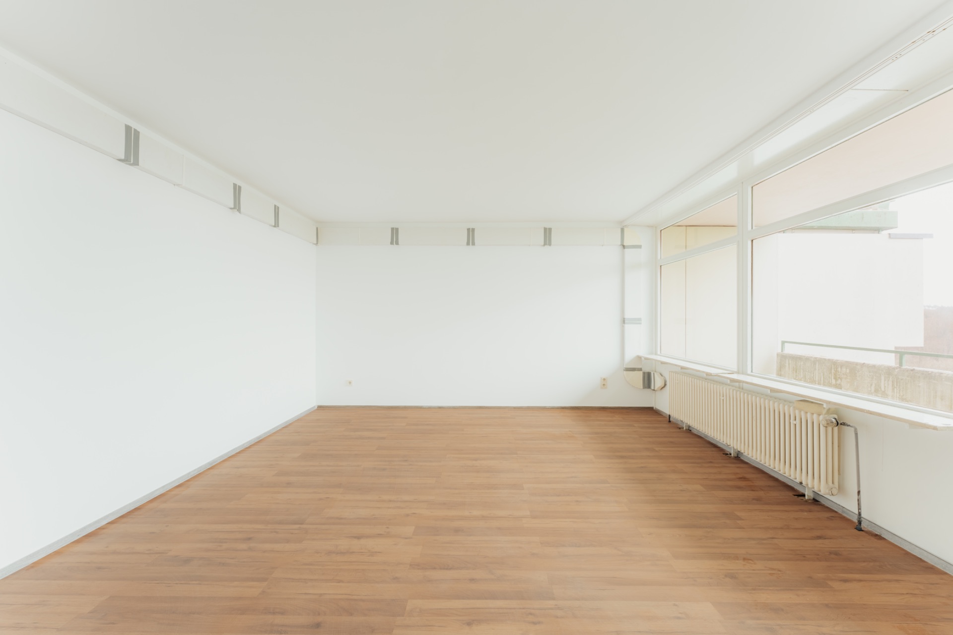 Leerer Raum mit weißer Wand, großen Fenstern und Holzfußboden.