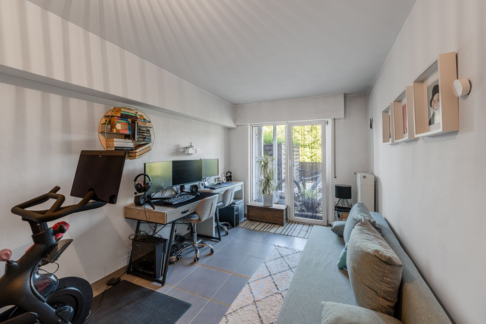 Wohnzimmer mit Sofa, Schreibtisch, zwei Monitoren, Heimtrainer und Zugang zur Terrasse.
