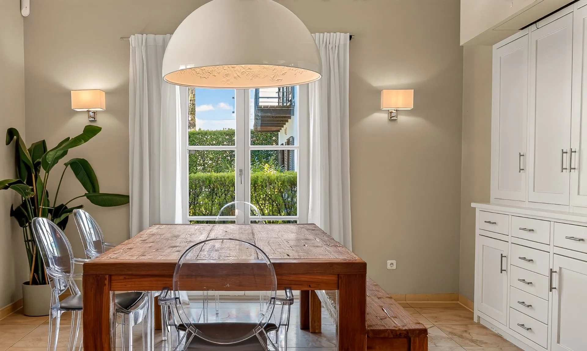 Esszimmer mit Holztisch, transparenten Stühlen, großer Lampe und Fenster mit weißen Vorhängen.
