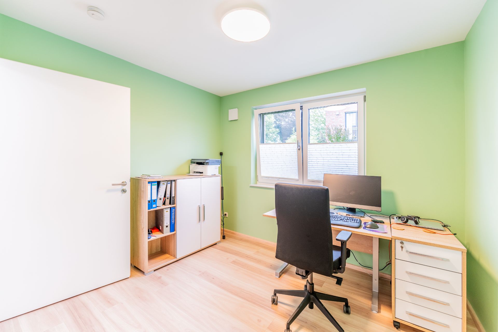 Helles Arbeitszimmer mit grün gestrichenen Wänden, Schreibtisch, Stuhl, Computer und Regal.