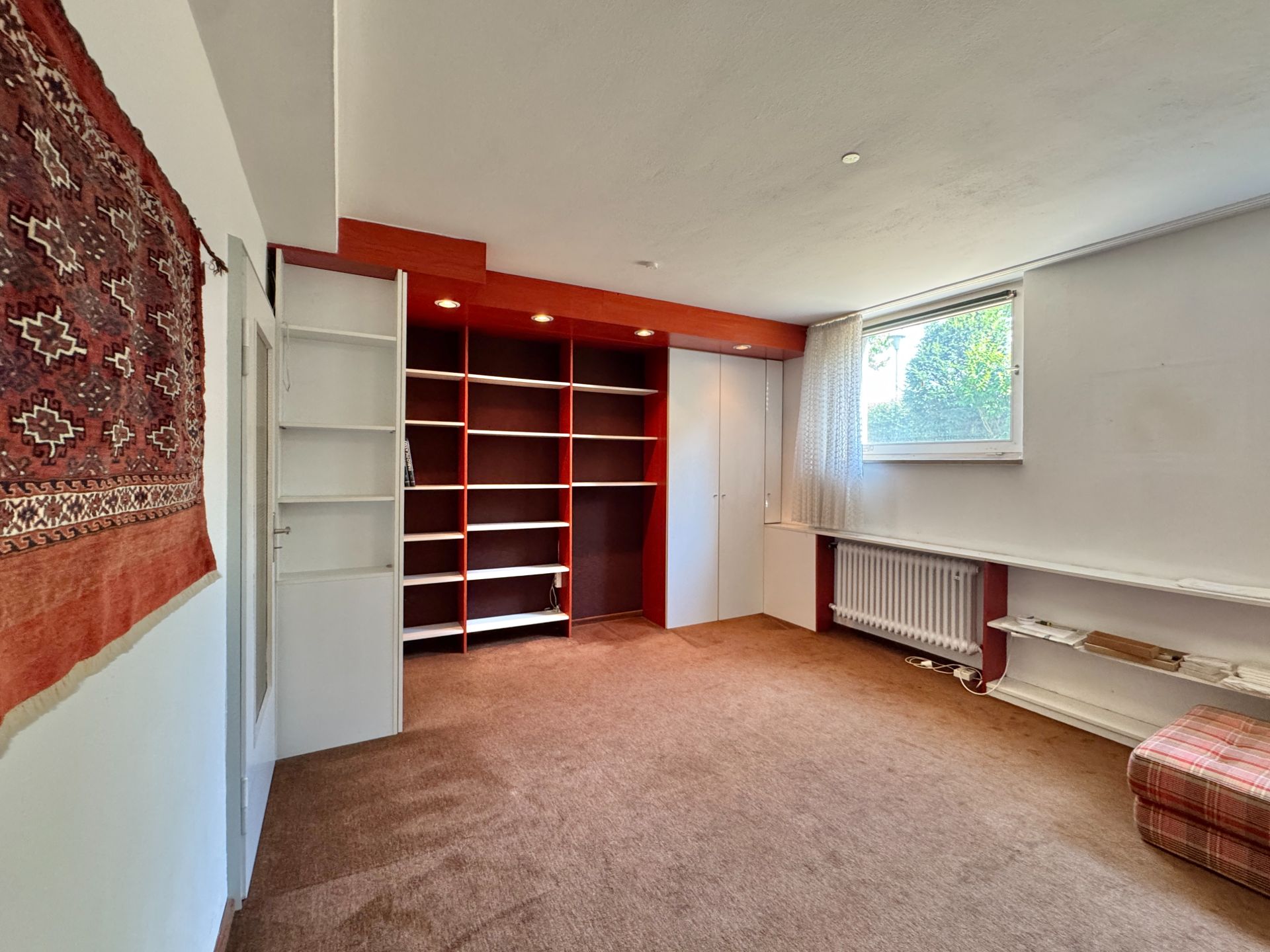Zimmer mit braunem Teppich, großem Regal, Wandteppich und Fenster mit Vorhang.