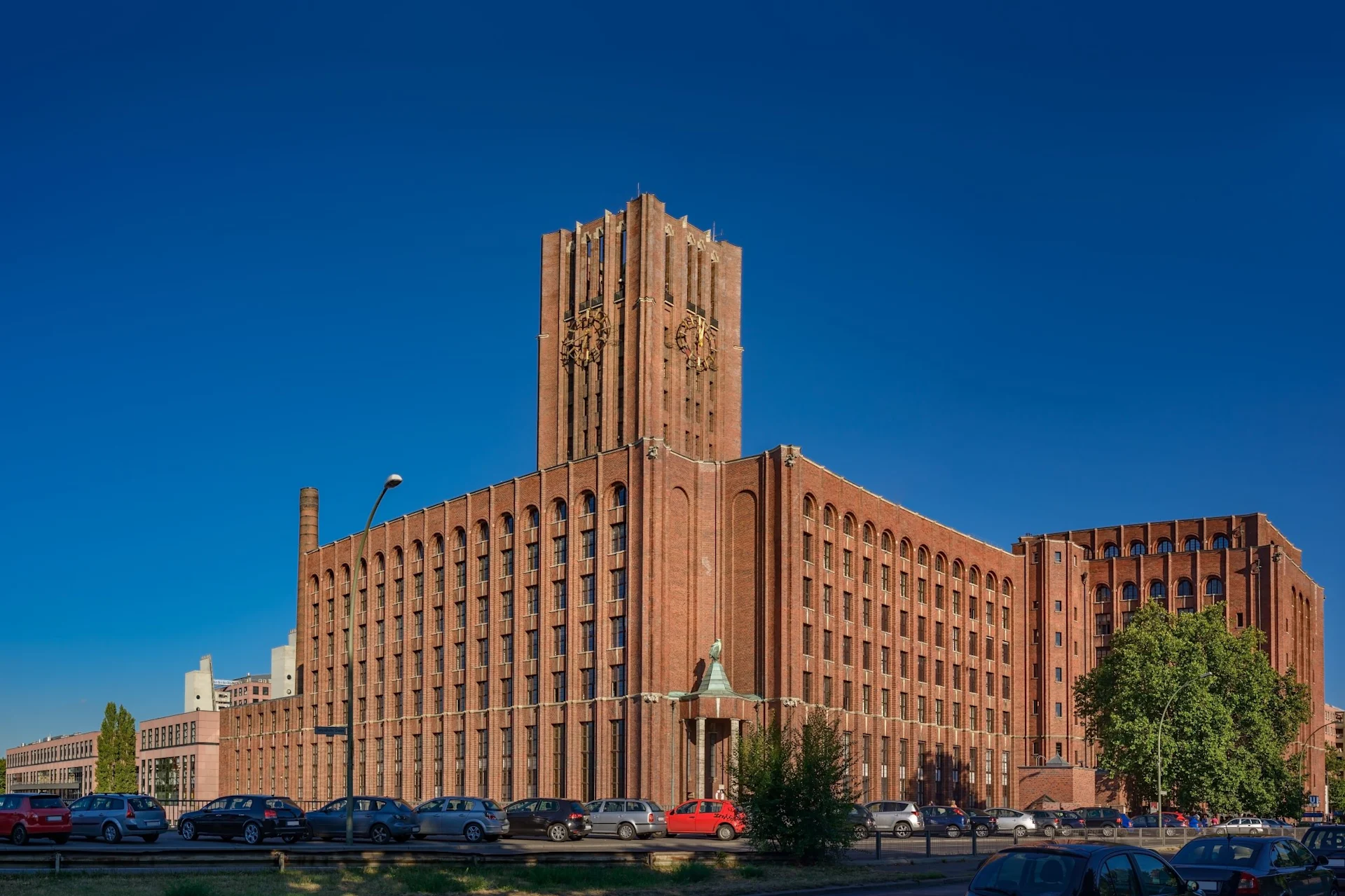 Großes Backsteingebäude mit Turm und vielen Fenstern, davor geparkte Autos und Bäume.
