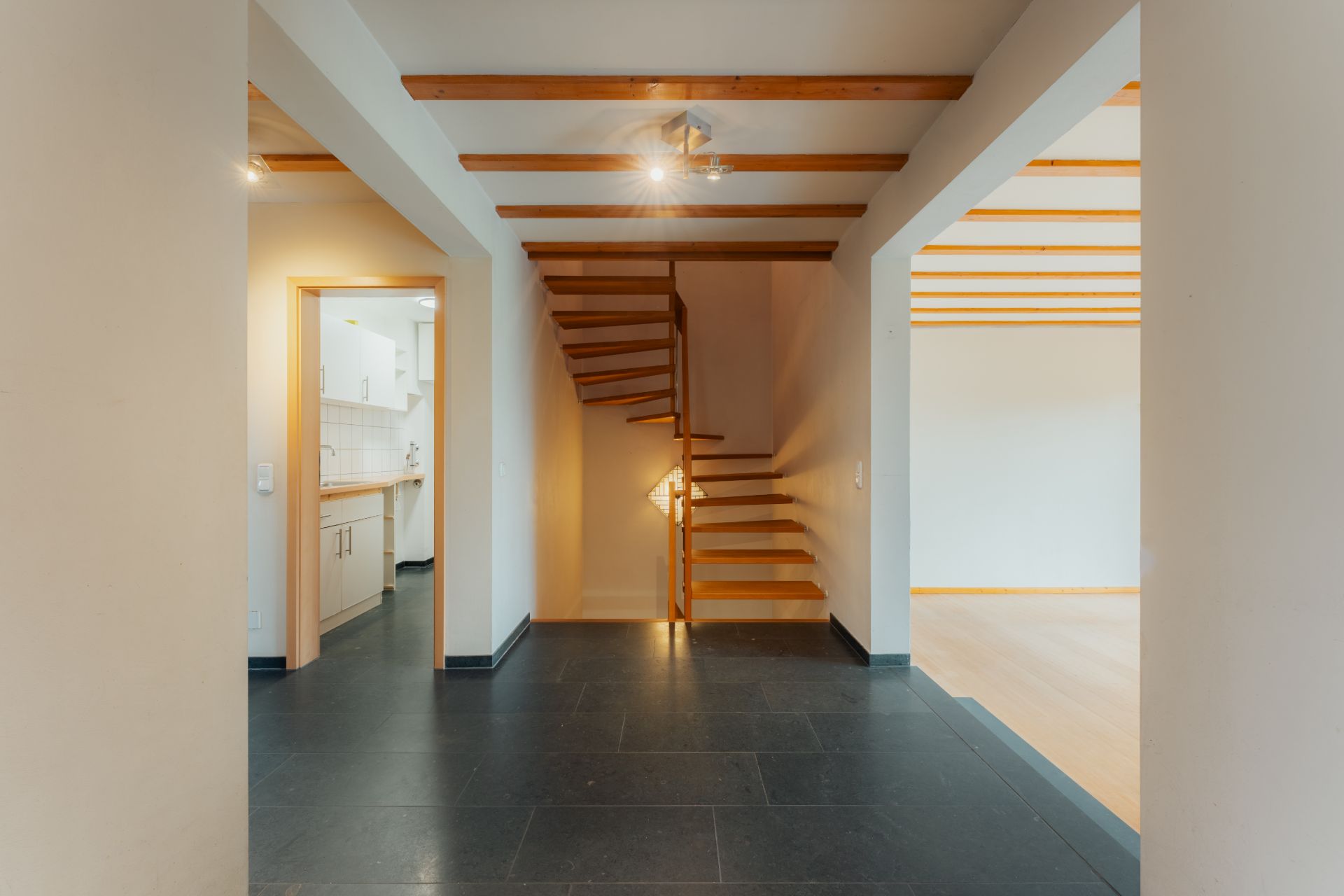 Flur mit schwarzem Fliesenboden, Holztreppe und Zugang zur Küche und Wohnzimmer.