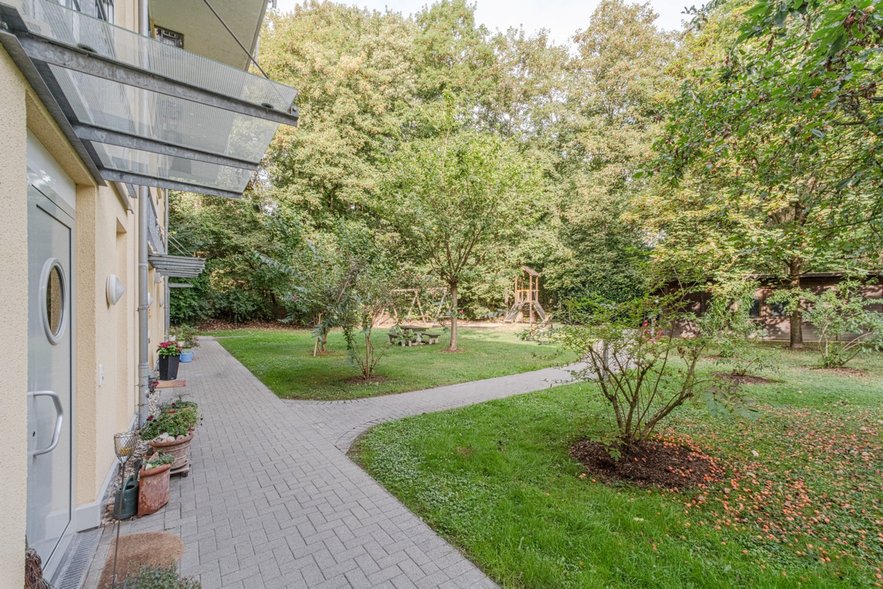 Gepflegter Garten mit Spielplatz, Wegen und Bäumen neben einem Wohngebäude.