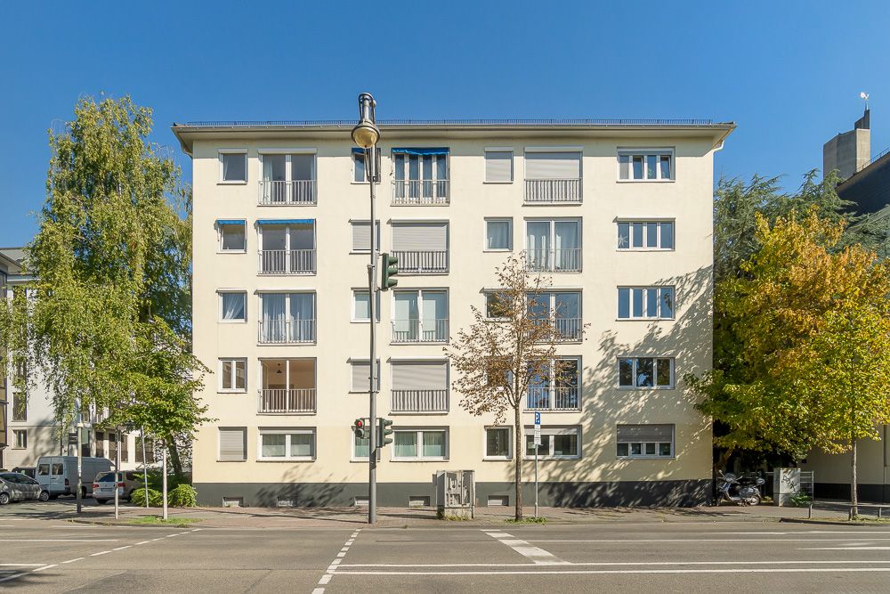 Vierstöckiges Wohngebäude mit mehreren Fenstern und Balkonen, Bäume davor und blaue Himmel.