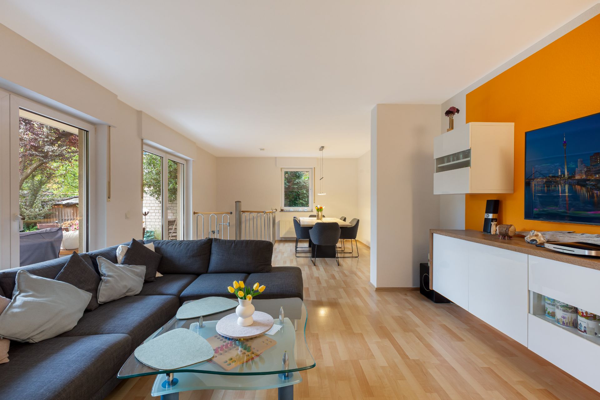 Modernes Wohnzimmer mit großem Sofa, Glastisch, Holzfußboden und Essbereich im Hintergrund.